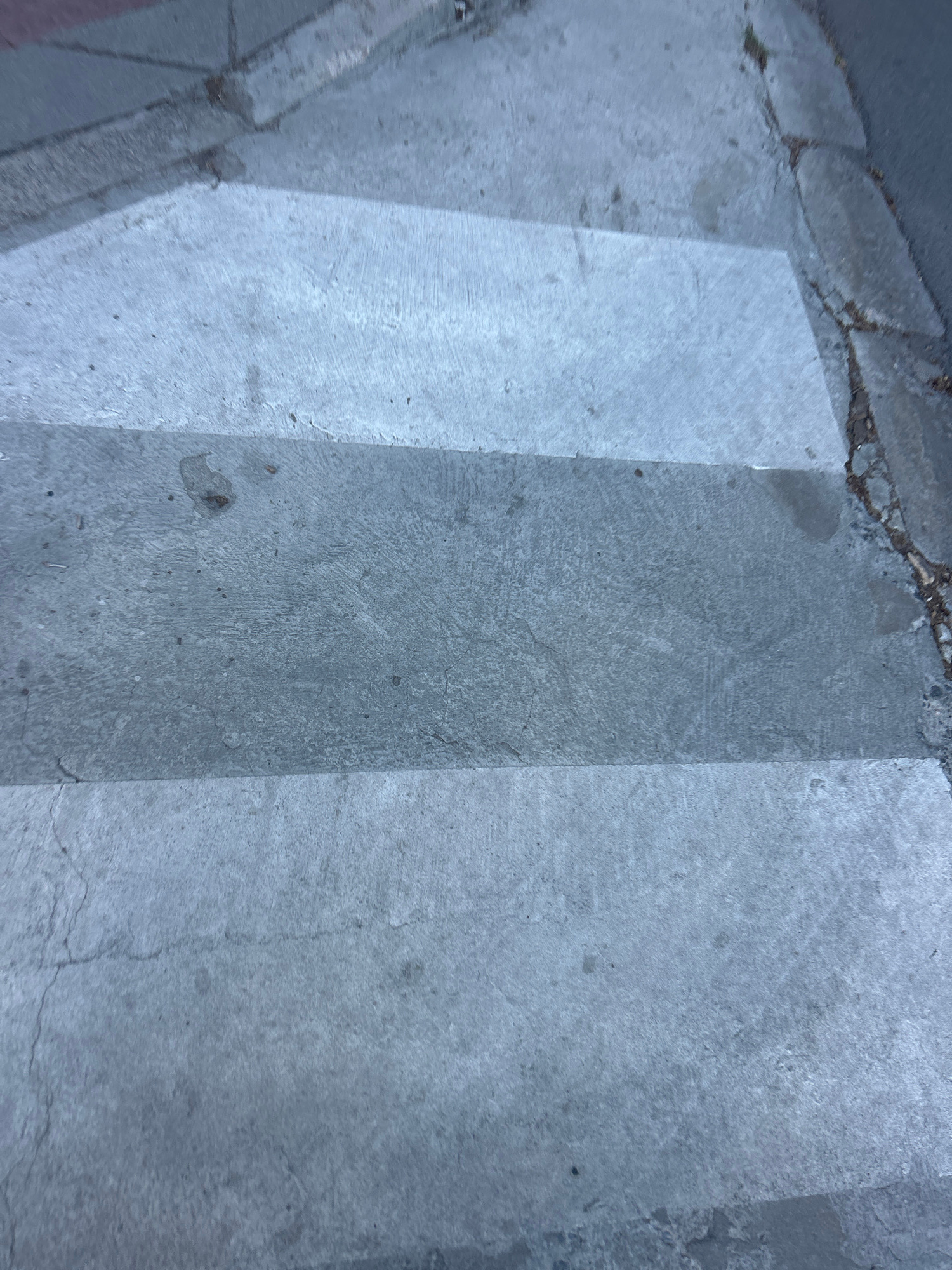 A high-angle, close-up shot shows a crosswalk with alternating light and dark grey concrete stripes. The surface is textured and shows signs of wear, with some cracks and small dark spots scattered across the pavement. A concrete curb with small green weeds growing in the cracks separates the crosswalk from an asphalt road on the right side of the frame. The scene is devoid of people or any discernible activity.