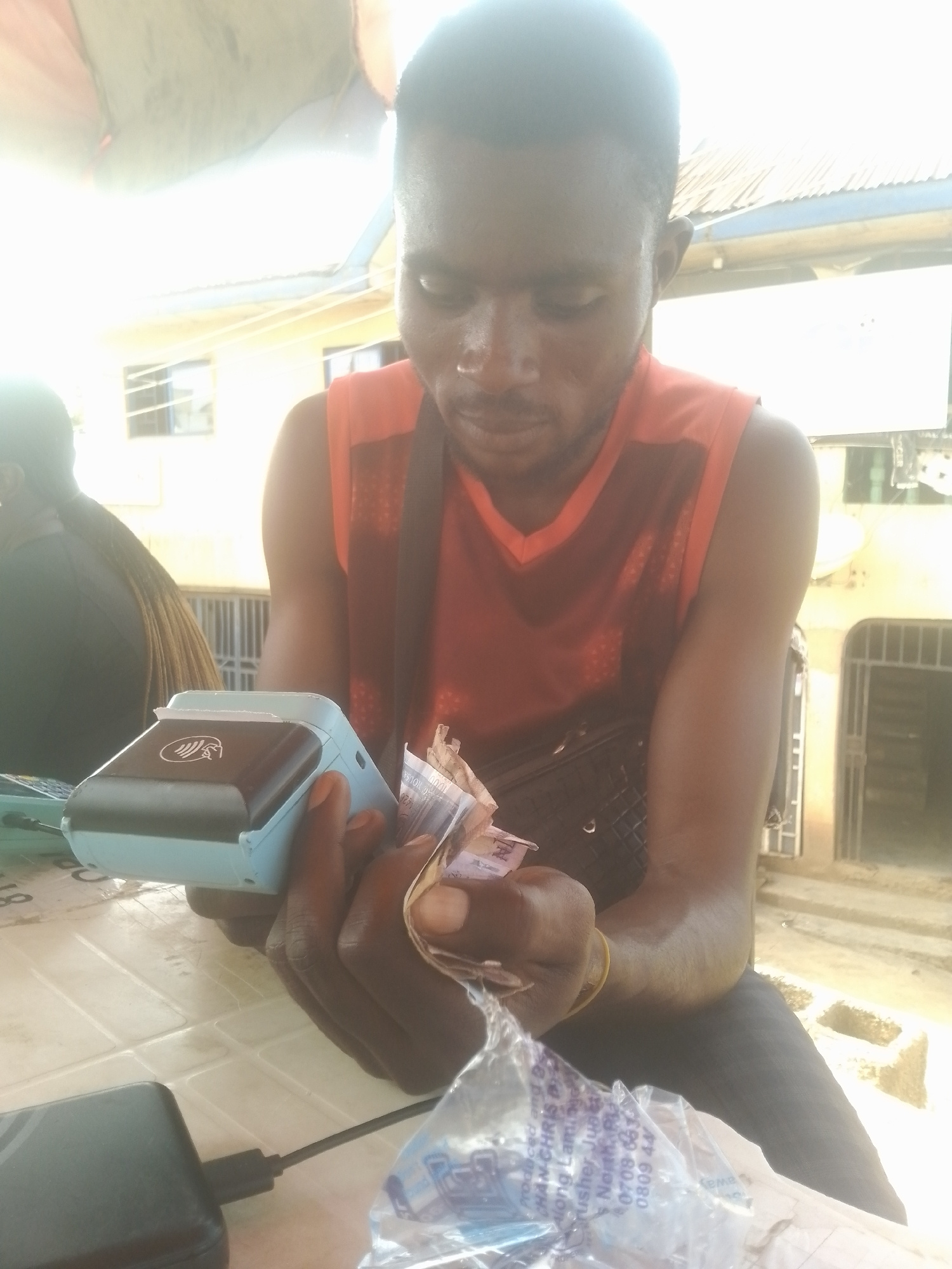 A medium shot, captured from a slightly low angle, shows a dark-skinned man in a red sleeveless shirt holding a portable point-of-sale device and money. The man is seated at a table outdoors. His gaze is directed downwards at the device and the bills in his hands. A black bag is slung across his chest, and a yellow wristband is visible on his left arm.
In the foreground, to the left of the POS device, a black power bank with a charging cable is on the table. To the right, a plastic wrapper with text is also visible on the table. In the background, a person with braided hair is partially visible on the left. The environment appears to be a street or open market with buildings and a corrugated metal roof in the distance. The lighting is bright, suggesting daytime.