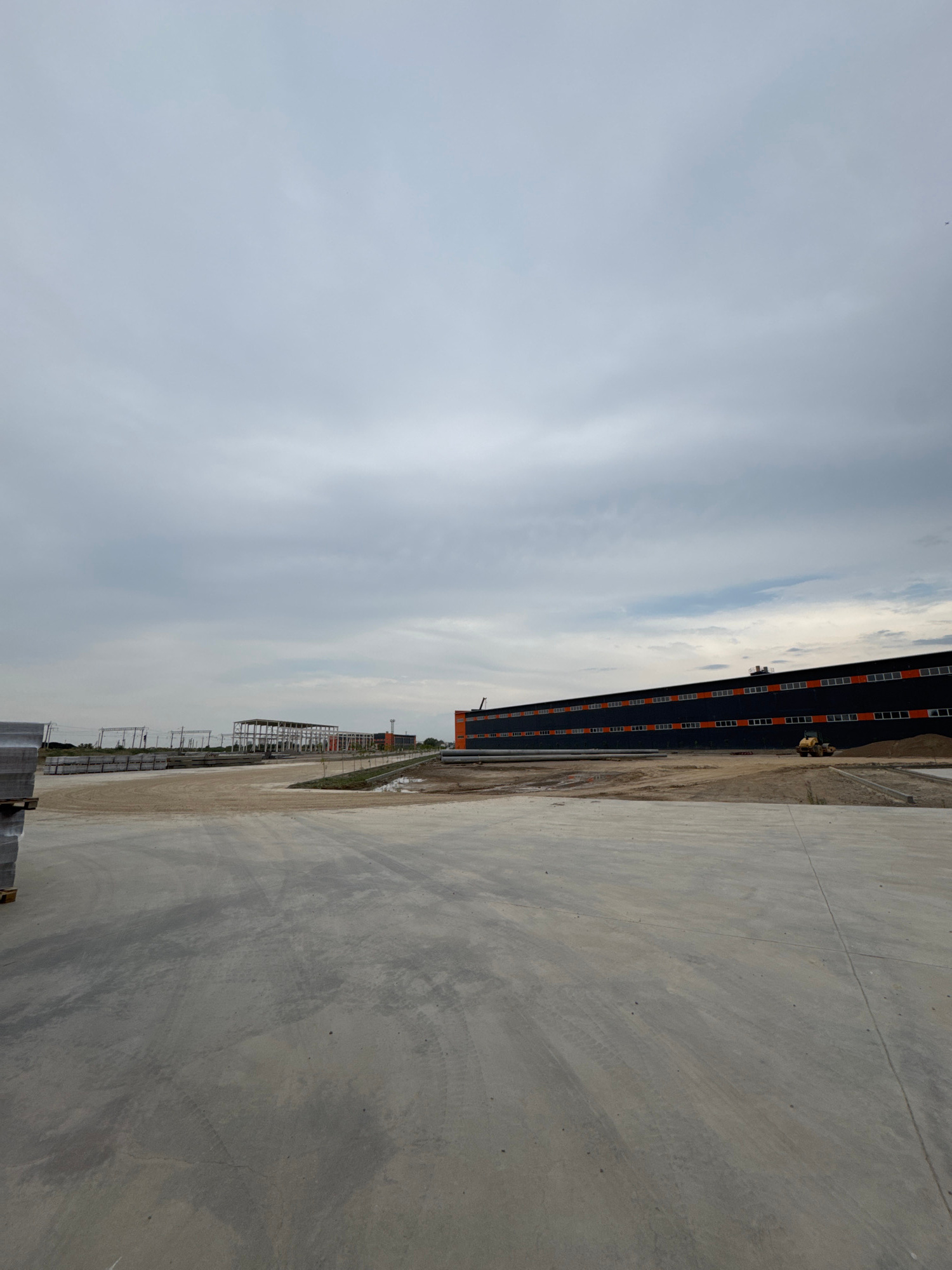 An outdoor, ground-level, wide-angle shot captures a construction site under a cloudy sky. In the foreground, a large, flat, grey concrete surface dominates the lower half of the frame, showing tire tracks from vehicles. To the left, a stack of grey concrete slabs is partially visible. In the mid-ground, an unfinished skeletal structure of a building is present, along with a long, dark building with orange rectangular window-like details. A yellow compact roller is positioned on the concrete in front of the dark building. The background consists of a distant treeline and more construction in the far distance. The scene appears to be located in Shamalgan, Kazakhstan, a developing industrial or commercial area. There are no people visibly present in the image.