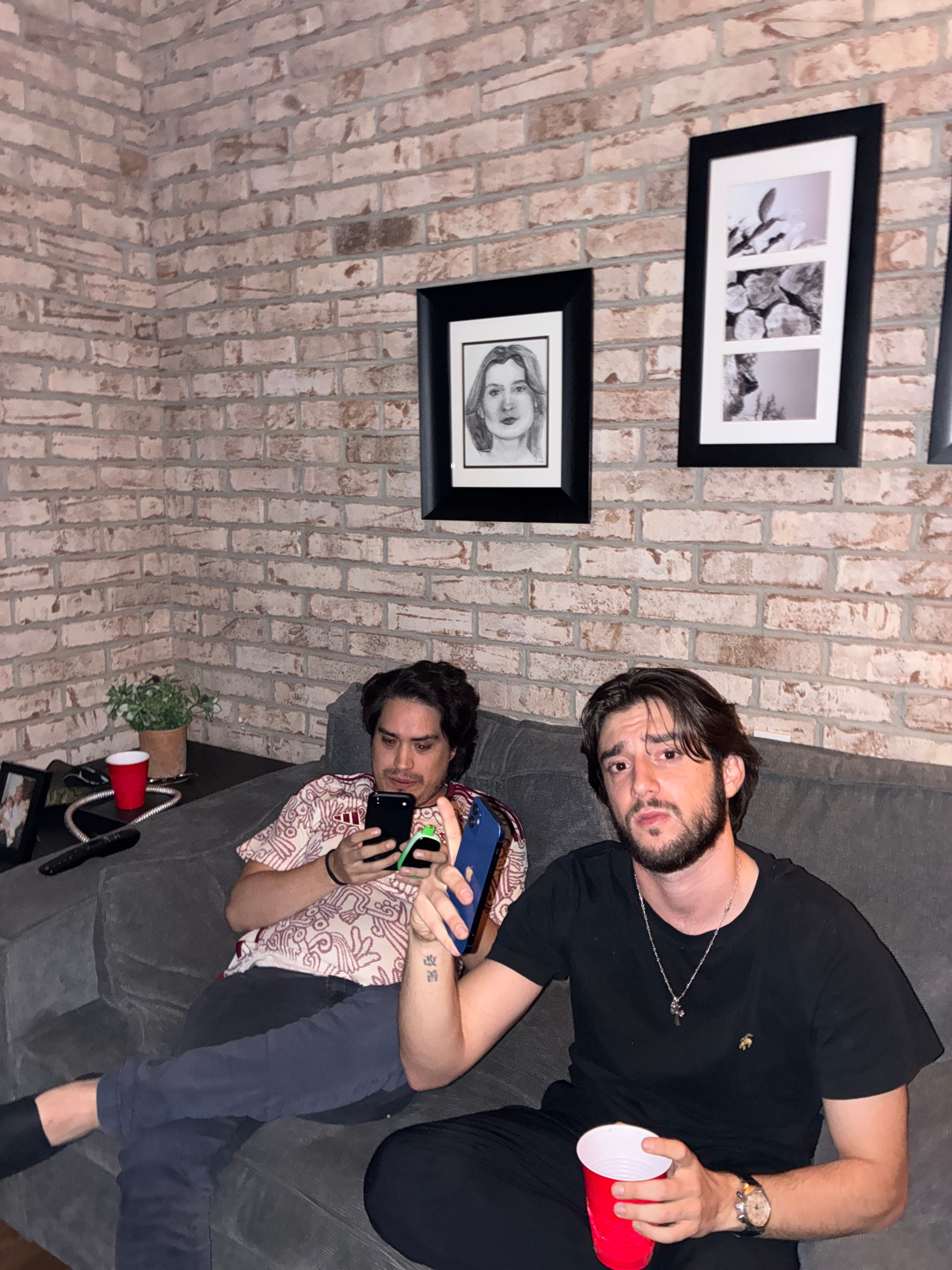 This media file features two individuals relaxing on a couch against a brick wall. The person on the left is lying down, looking towards the camera with a relaxed expression. They are wearing a black t-shirt, a silver necklace with a pendant, and a watch on their left wrist. They are holding a red disposable cup in their right hand.
The person on the right is also lying down on the couch, facing away from the camera. They are wearing a patterned shirt and dark pants. They are holding two smartphones, one in each hand, and appear to be engaged with them. A tattoo is visible on their left arm.
The scene appears to be indoors, with a brick wall providing a textured backdrop. On the wall, there are two framed pictures. The upper frame contains a triptych of black and white photographs, possibly of nature or abstract subjects. The lower frame displays a black and white portrait of a woman.
On the couch, near the person on the right, there is a small potted plant and another red disposable cup. The lighting suggests it might be evening or indoors with artificial lighting. The overall mood is casual and relaxed. There is no visible text in the image. The setting suggests a home or casual gathering space.