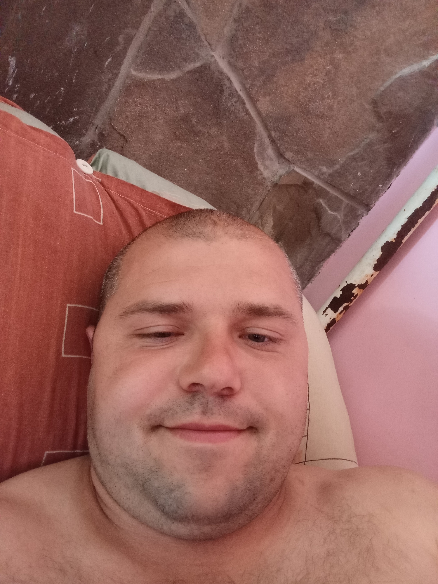 A man is lying on a pillow with a reddish-orange pillowcase. He is wearing a light-colored shirt, and the top of his chest is visible. The background appears to be a rustic stone wall and a pink surface with a rusty pipe. The man's head is tilted slightly to his left, and his left eye is closed or partially closed, giving him a winking appearance. The lighting is dim. The scene is indoors, likely in a bedroom or a similarly private space. The city and country are unknown.