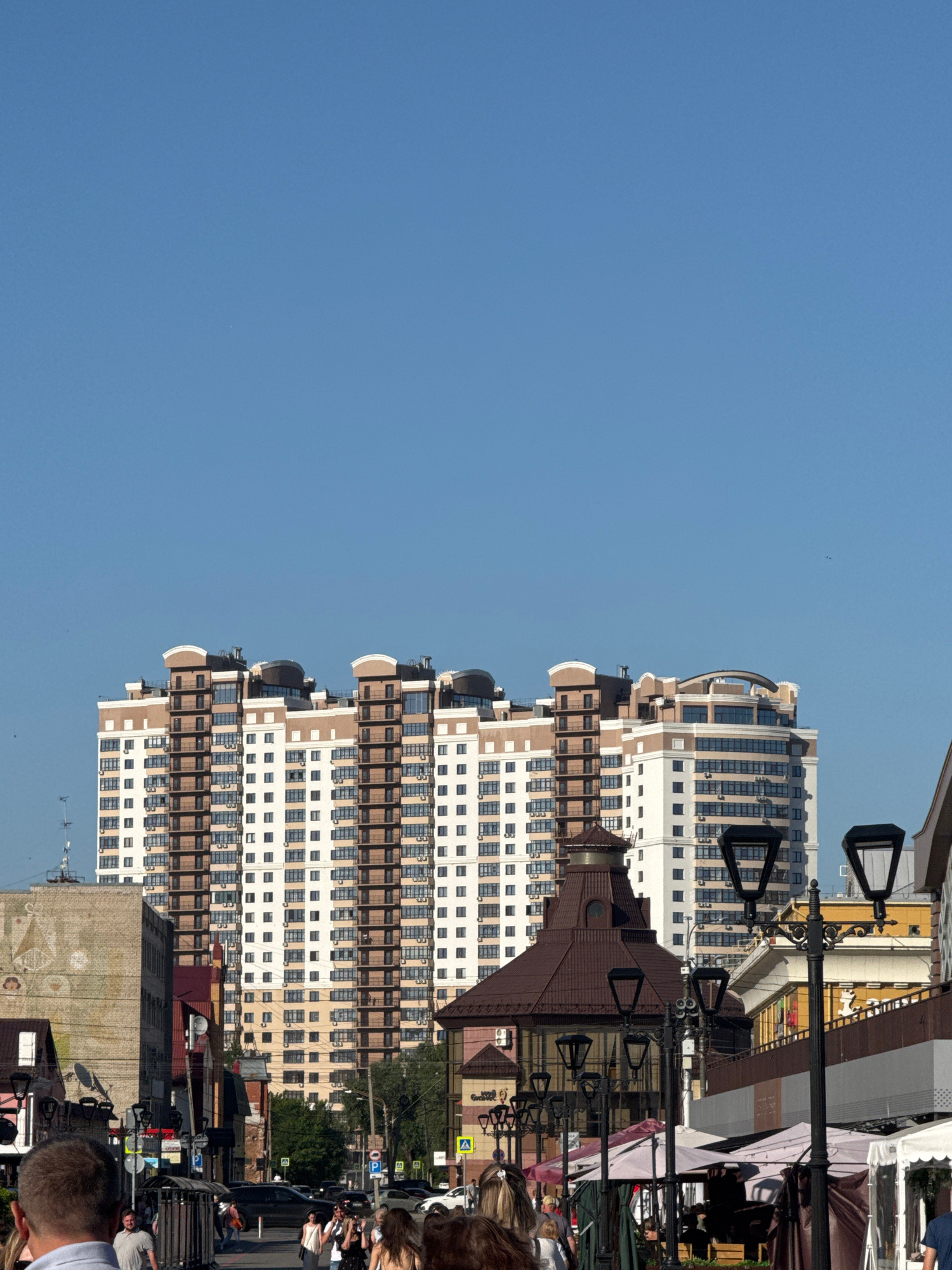 The image captures a street scene in Barnaul, Russia, dominated by a large, modern apartment building with a beige and white facade and numerous balconies. In the foreground, a street lined with market stalls and lampposts is visible. Several people are walking along the street, some in the foreground and others further back. The sky is clear and blue.