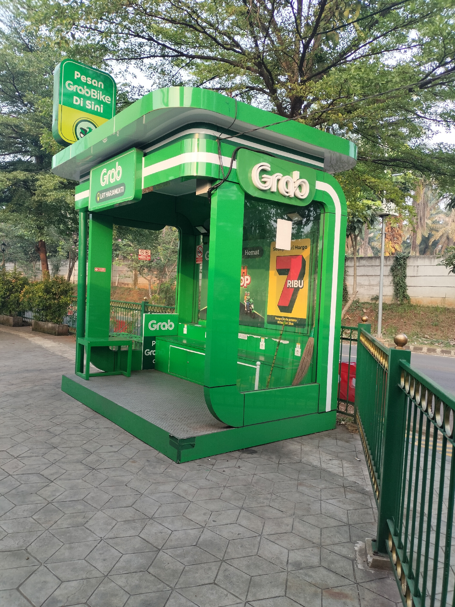 A green GrabBike station is situated on a patterned concrete sidewalk adjacent to a road in Depok, Indonesia. The station is a small, open-sided structure with a green roof and white trim. A prominent green sign on the roof reads "Pesan GrabBike Di Sini" (Order GrabBike Here) in white text with a yellow circular graphic below. Below the roof, there are two smaller green signs with the Grab logo.
Inside the station, there are two benches and a small table on the left side. The right side of the station features a large poster with the Grab logo and text that appears to be promotional, including the number "7" and "RIBU" (thousand), suggesting a price or offer. The interior also contains a broom and a red object visible through the glass on the right.
The environment surrounding the station includes trees with lush green foliage, a metal railing along the sidewalk, and a concrete wall in the background. The sidewalk has a repeating geometric pattern of interconnected cubes. The overall lighting suggests it is daytime, possibly late afternoon or early morning given the soft light. There are no people visible in the image.