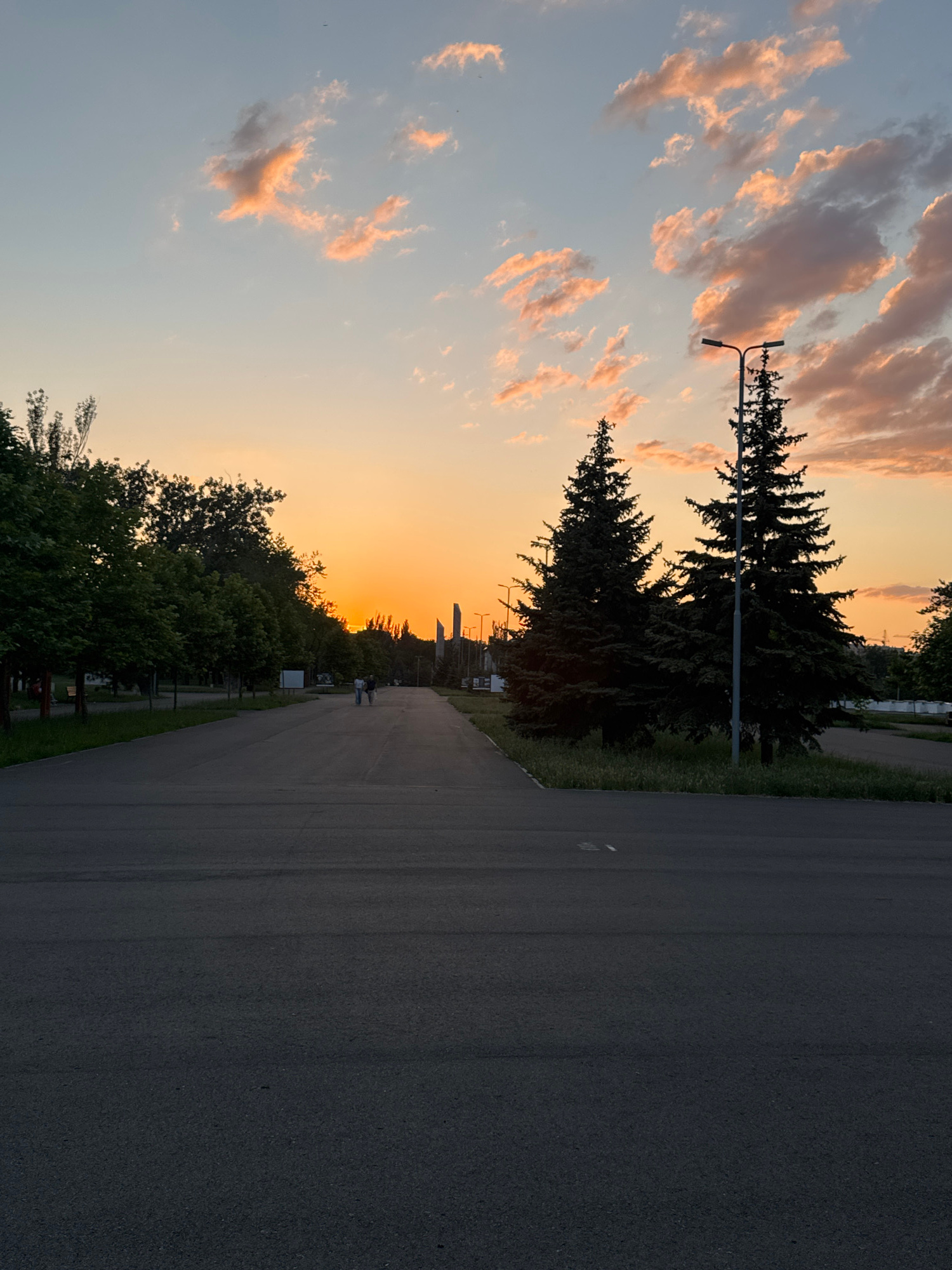 The image depicts a pathway lined with trees on the left and two large pine trees on the right, with a street light between them. The sky is filled with clouds illuminated by a sunset. In the distance, two people are walking on the pathway. The environment appears to be a park or public area. There are structures in the far distance, suggesting an urban setting. The scene is set in Mariupol, Ukraine.