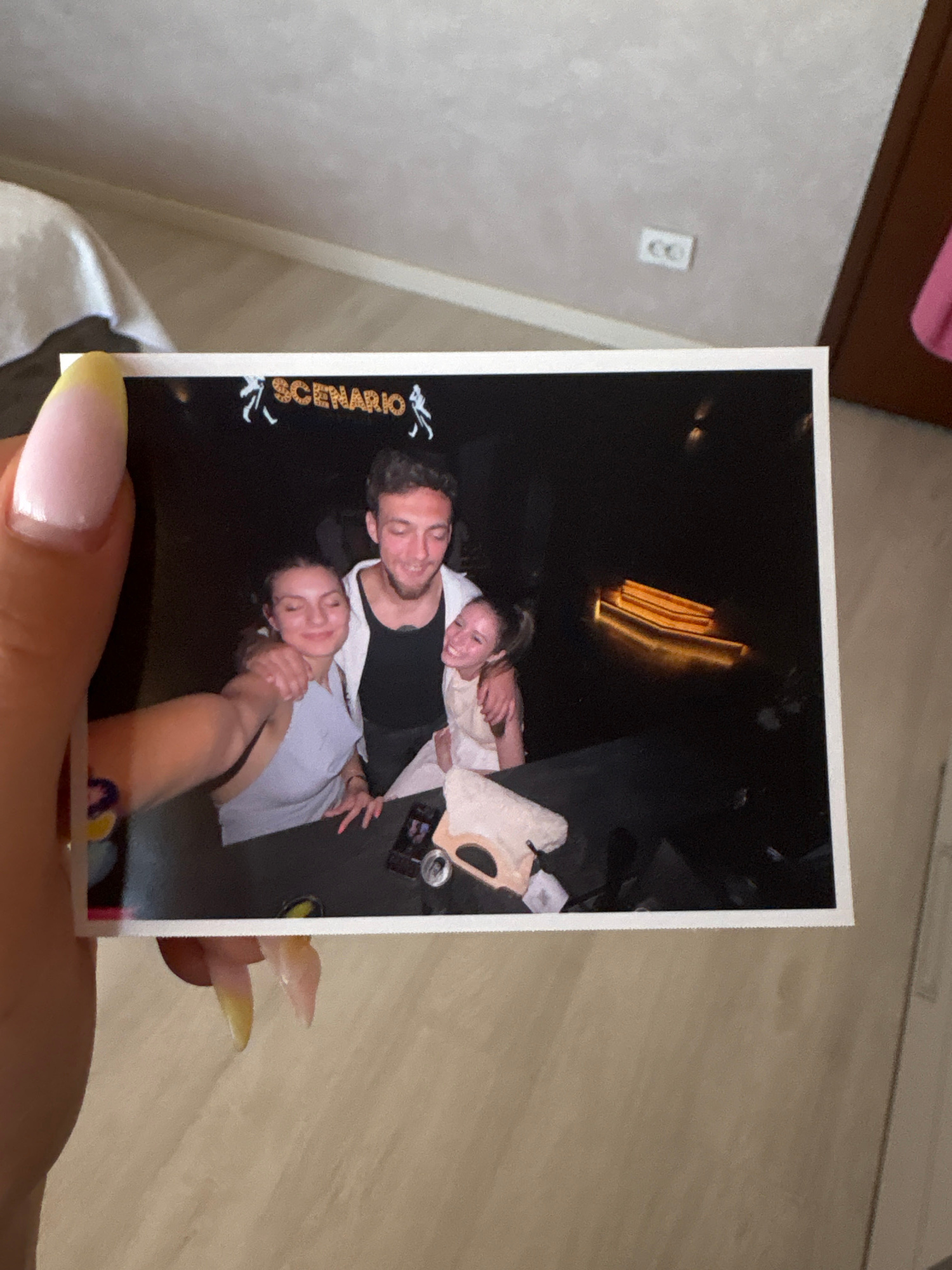 The image shows a photograph of four young adults posing together. Two women and two men are visible. The man in the center is wearing a white hoodie over a black tank top and has his arm around one of the women. The woman next to him is wearing a light-colored top and has her head tilted back with her eyes closed. Another woman is in the foreground on the left, with her arm around the man. A fourth person, partially obscured, is behind the man. The photograph appears to have been taken in a dimly lit environment, possibly a club or a party, indicated by the glowing elements in the background and the overall dark setting. The word "SCENARIO" is visible in glittery lettering at the top of the photograph, likely the name of the venue or event. The photograph is being held up by a person's hand with prominent fingernails, suggesting it's a physical print. There is no information about the location in Bulgarovo, Bulgaria.