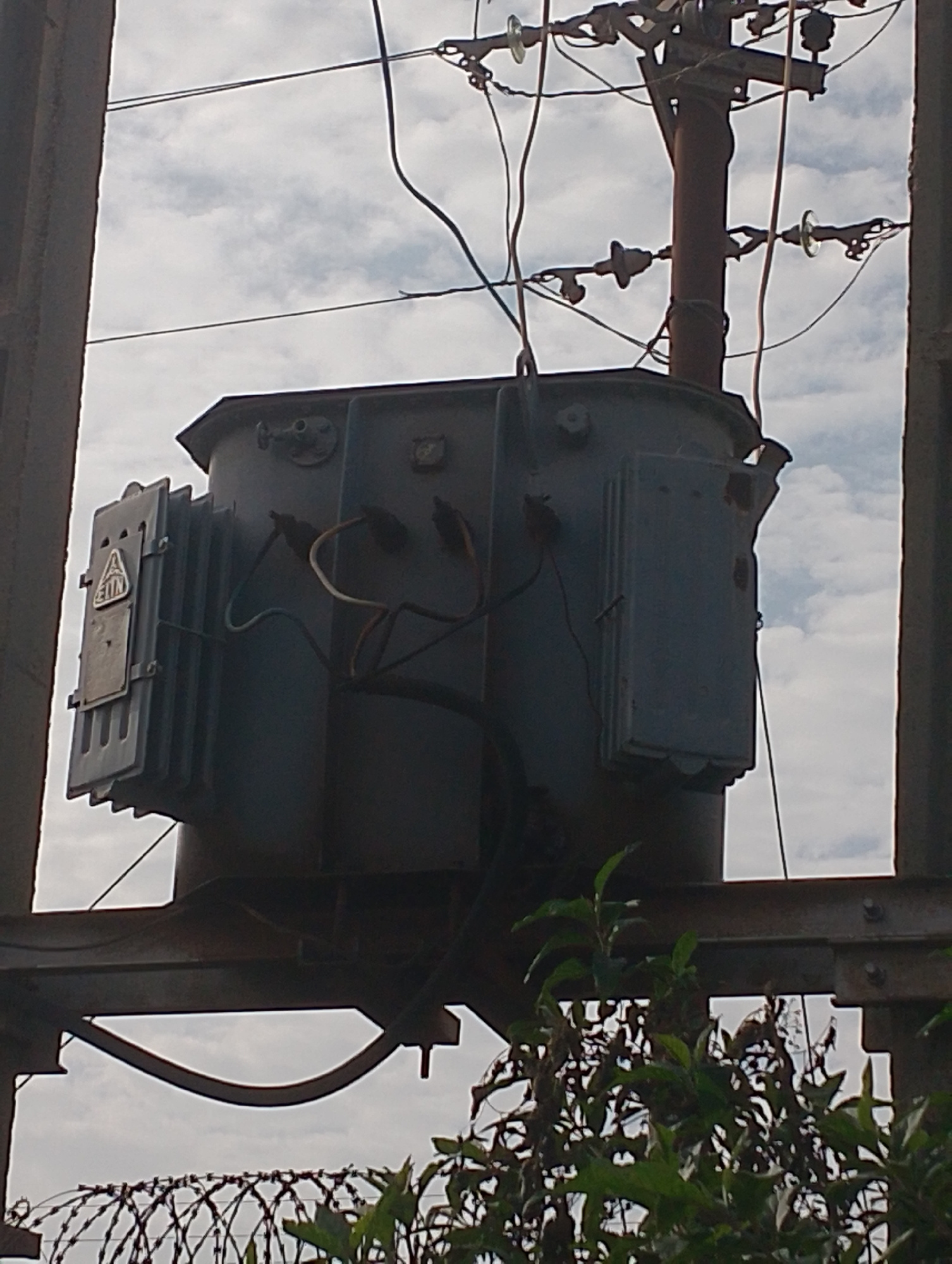 This image depicts an electrical transformer mounted on a metal support structure, situated outdoors. The transformer is a large, gray, rectangular unit with various electrical components and wiring attached. It appears to be part of the city's electrical infrastructure. In the background, a pole with insulators and overhead power lines is visible against a cloudy sky. In the foreground, there are green leafy plants and barbed wire, suggesting the transformer might be in a secured area or an industrial zone. The overall scene is a snapshot of urban utility infrastructure. The location is identified as Jos, Nigeria.