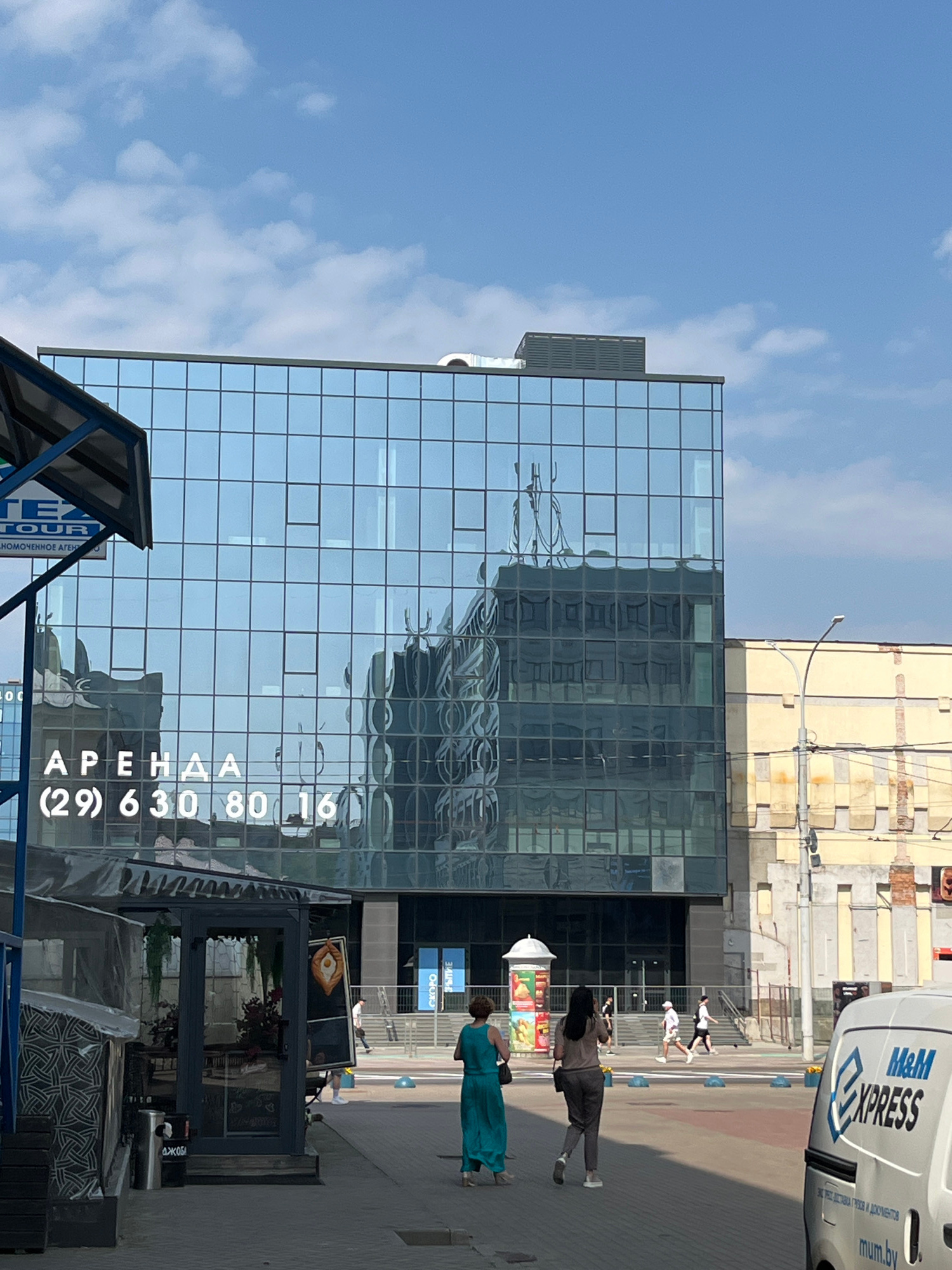 The image shows a street scene in Minsk, Belarus, featuring a modern glass-fronted building with the words "АРЕНДА" (RENT) and a phone number "(29) 630 80 16" displayed prominently. The building's facade reflects the sky and surrounding structures, including an older yellow building with visible brickwork. In the foreground, several people are walking on a paved area. One woman in a teal dress is walking away from the camera, and another woman in gray pants and a brown top is walking in the same direction. A white van with "M&M EXPRESS" branding is parked on the right side of the image. The environment appears to be an urban commercial area under a clear blue sky with some clouds.