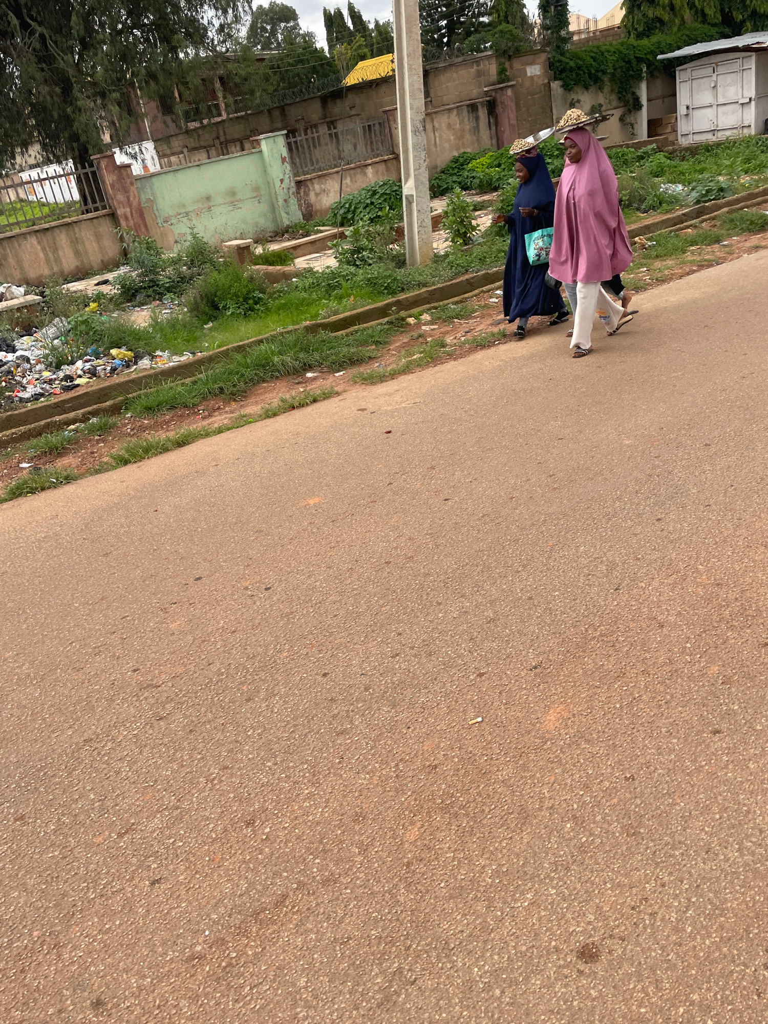 In Jos, Nigeria, two women walk on a paved road. The woman on the right wears a pink hijab and white pants, carrying a flat tray balanced on her head. The woman on the left wears a dark blue hijab and a bag with a patterned design slung over her shoulder. Behind them, a roadside ditch is filled with accumulated garbage and debris, bordered by a low concrete wall and overgrown grass. In the background, a fence separates the road from a property with a green wall, trees, and buildings. A white metal container is visible on the right side of the background. The sky is overcast.