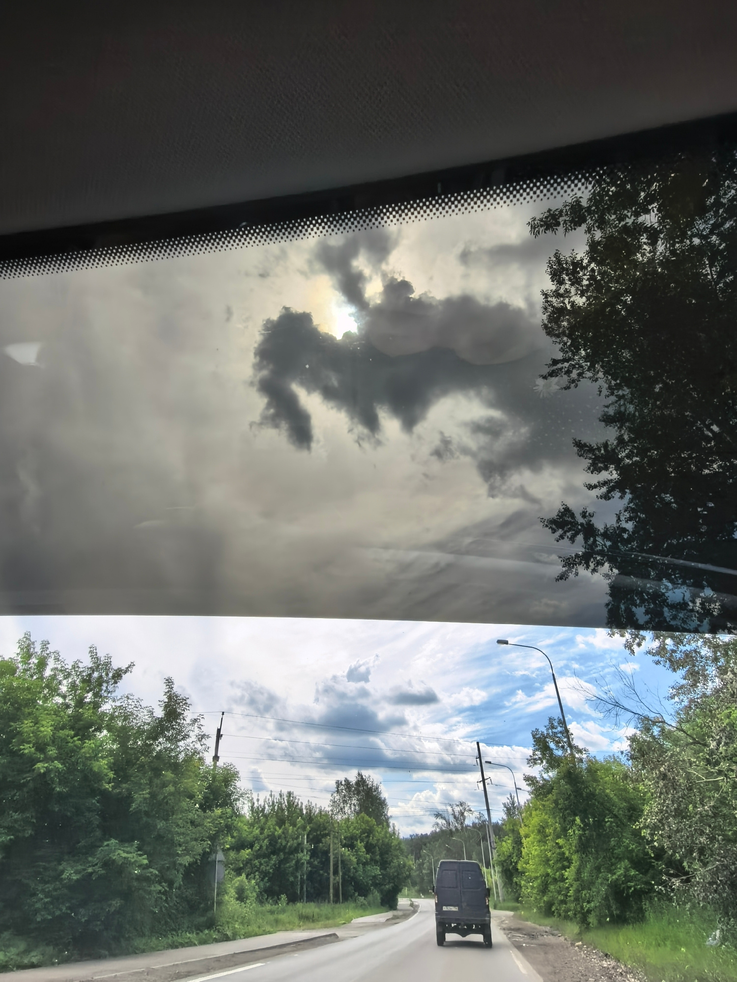 The image is a split composition showing two scenes. The top half, viewed through a windshield, depicts a cloudy sky with the sun partially obscured by dark clouds, creating a dramatic effect. The bottom half shows a two-lane road in a rural or suburban area, bordered by lush green trees on both sides. A dark-colored van is driving away from the viewer on the right lane. Power lines and streetlights are visible along the road. The scene appears to be captured during daylight hours. The location is not specified in the prompt.