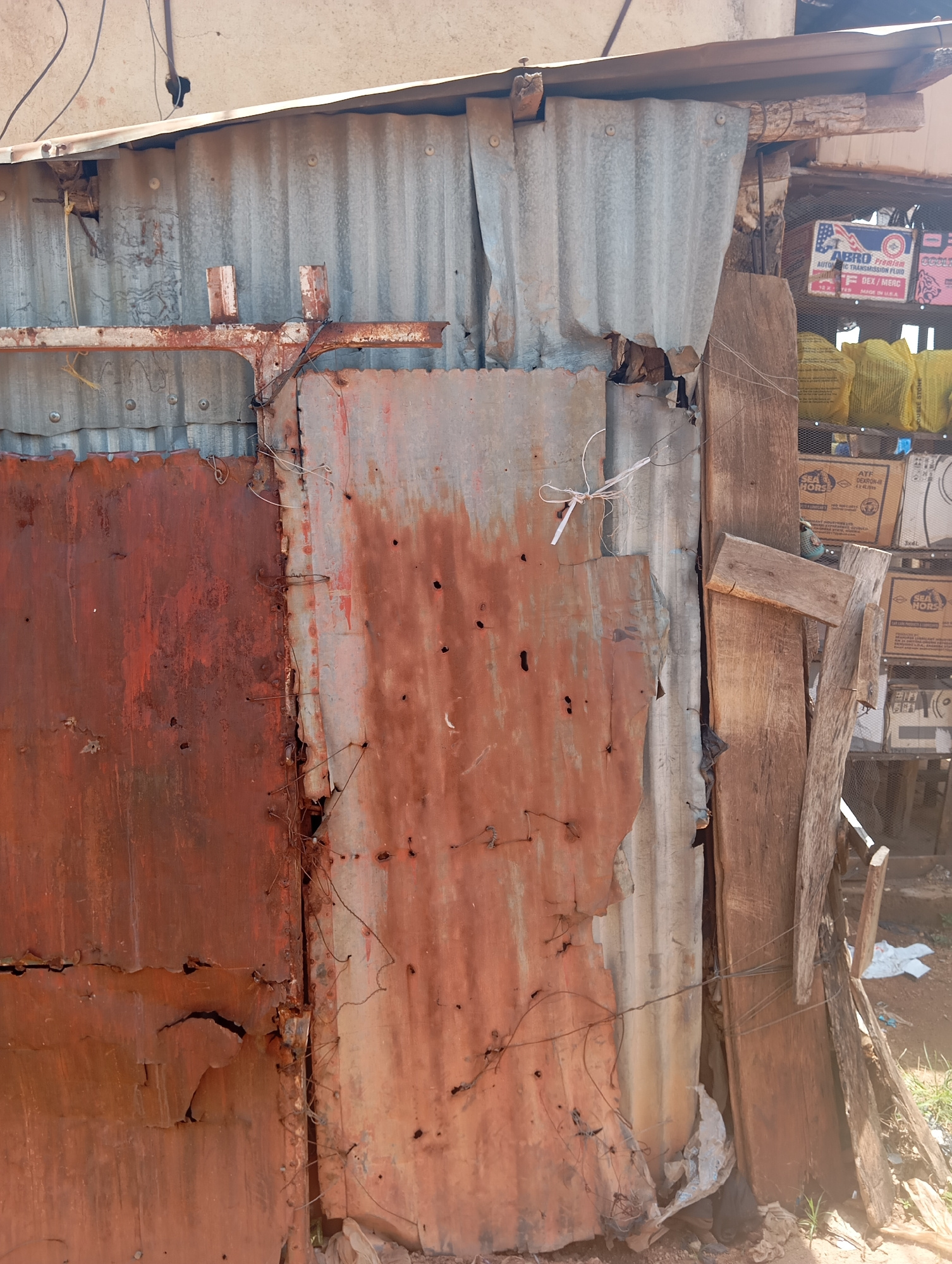 This image depicts a close-up view of a dilapidated, rustic structure, likely a shop or stall, in Jos, Nigeria. The primary subjects are the materials forming the structure: corrugated metal sheets, some heavily rusted and weathered, others appearing more galvanized. These sheets are in disrepair, with holes, tears, and visible seams held together by wire and nails. Wooden planks are also incorporated into the construction, adding to the makeshift appearance.
Behind the metal sheeting, shelves are visible, stocked with various items. Among these are boxes of "ABRO Automatic Transmission Fluid" and other products. Yellowish bags, possibly containing goods for sale, are also present. The ground appears to be dirt and scattered debris, suggesting an outdoor or semi-outdoor environment.
There is no discernible activity or event taking place; the scene is static, focusing on the texture and decay of the building materials. The lighting suggests it is daytime, with harsh shadows indicating bright sunlight. The overall impression is one of poverty or a struggling commercial enterprise, characterized by the use of salvaged and aged materials. The visible text provides brand names and product details for items being sold. The setting is clearly identifiable as Jos, Nigeria, adding a specific geographical context to the scene of makeshift commerce.