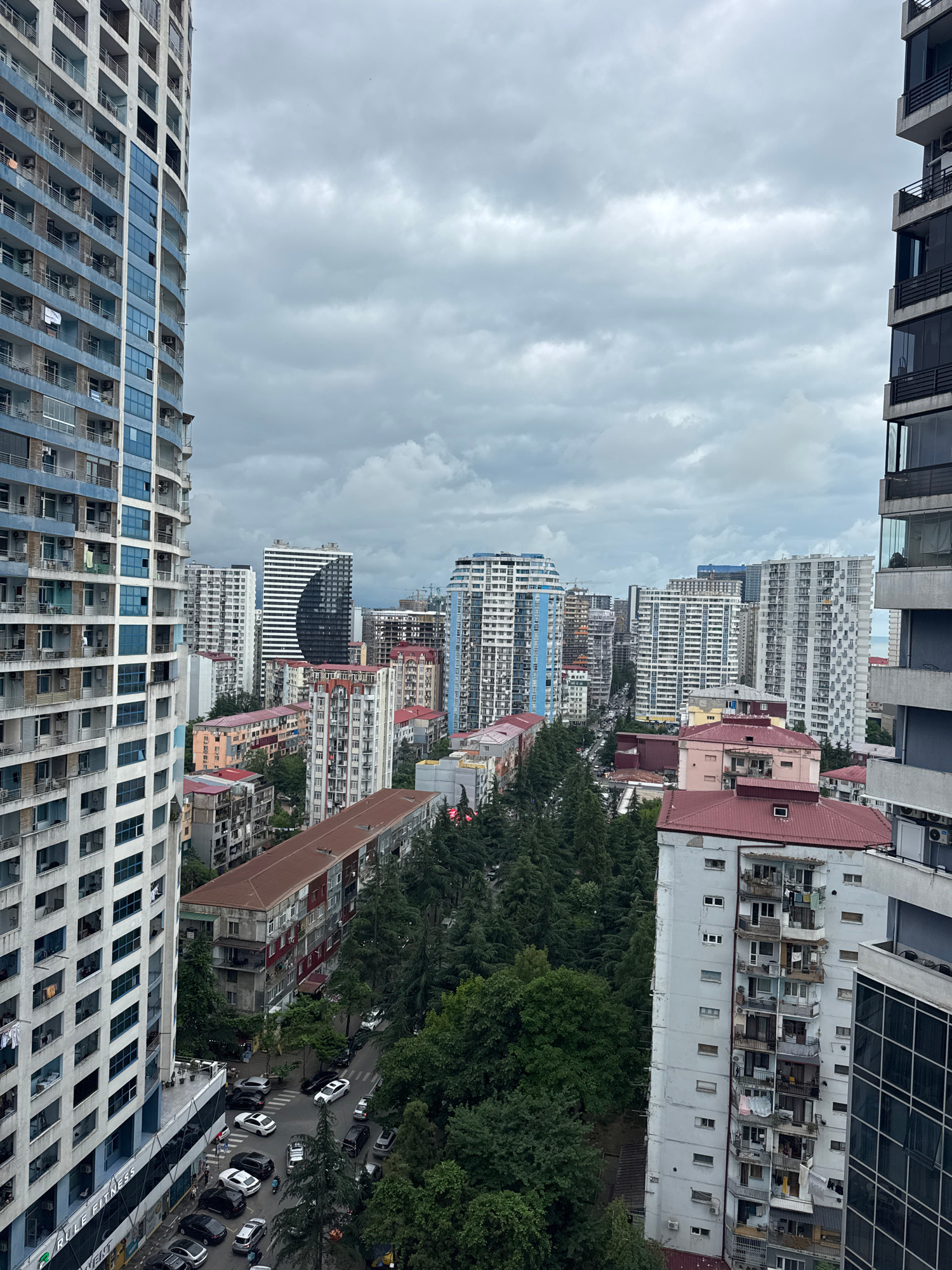 The image is a high-angle, outdoor view of a dense urban environment, likely in Batumi, Georgia, characterized by numerous multi-story apartment buildings and residential blocks. The architecture varies, with some modern, sleek high-rises featuring balconies and glass facades, and older, more weathered buildings with visible air conditioning units and laundry hanging from balconies. A street runs through the middle ground, lined with parked cars of various colors and models, suggesting a residential area. Lush green trees, primarily conifers, are interspersed between the buildings, creating a contrast between the urban structures and the natural elements. The sky is overcast with heavy, gray clouds, indicating a cloudy or potentially rainy day. The overall impression is one of a bustling, developed city with a mix of old and new constructions, set against a backdrop of a temperate, possibly coastal, climate.