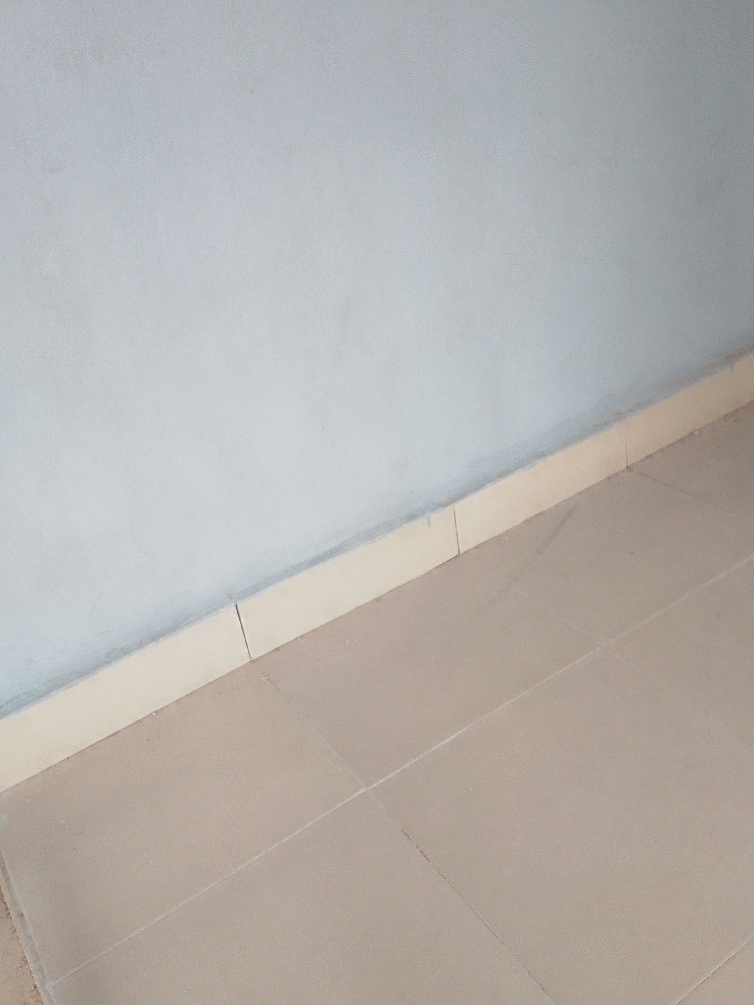 The image shows a corner of a room with a light blue wall meeting a tiled floor. A beige baseboard runs along the junction of the wall and floor. The floor is made of square, tan tiles with grout lines. The lighting suggests an indoor environment. There are no people or any discernible objects in the scene other than the architectural elements of the room. The image is likely a photograph taken from a fixed perspective, possibly for documentation or real estate purposes. The location is given as Unknown, Unknown.