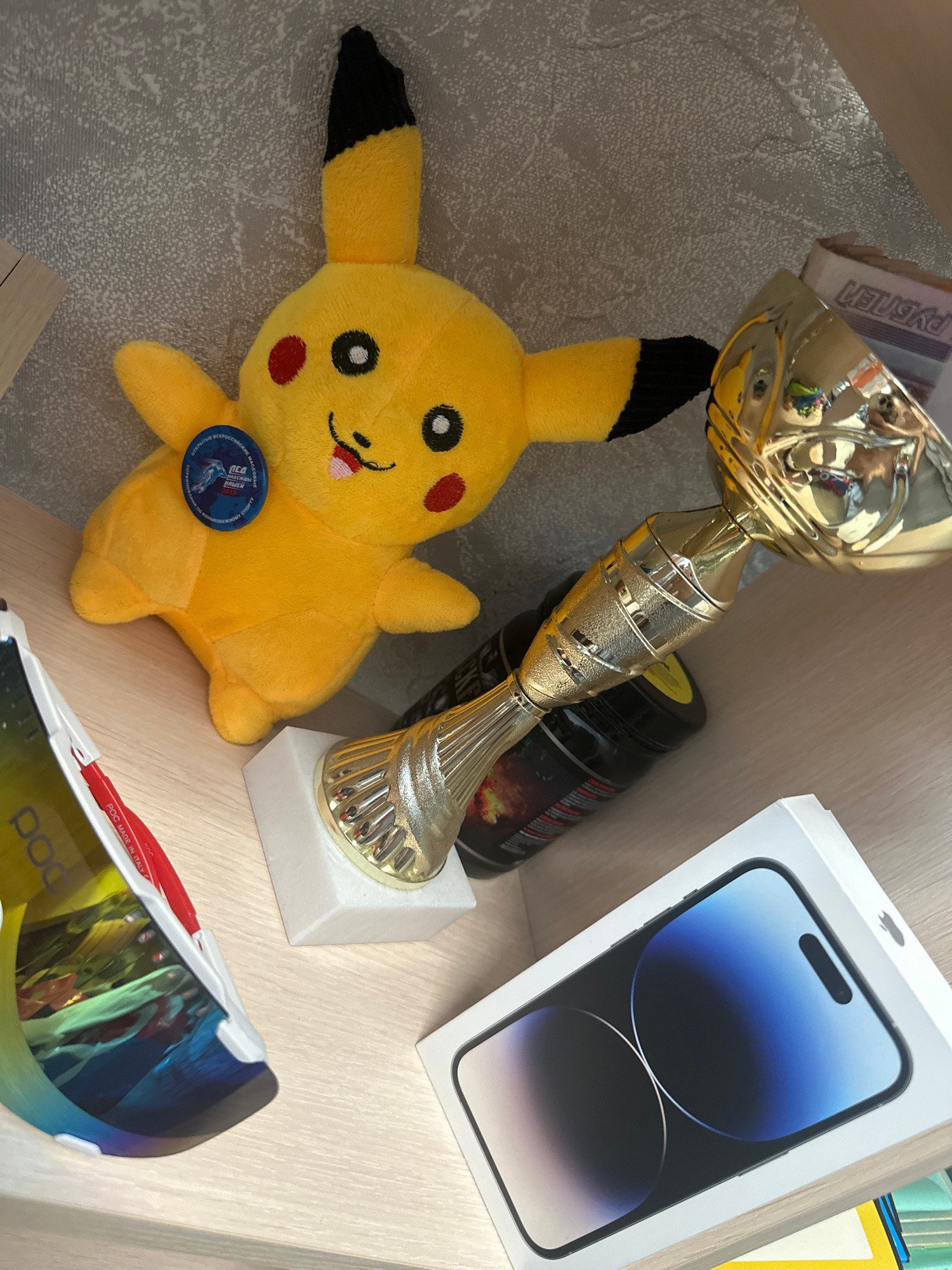 An indoor, eye-level, medium shot shows several objects arranged on a light-colored wooden shelf, possibly within a home or office setting. The background consists of a textured, light gray surface.
In the foreground and center-left, a yellow plush toy resembling the character Pikachu is positioned on its back. It has black-tipped ears, red circular cheeks, black eyes, and a smiling mouth with a red tongue. A small, circular blue badge with text and an image is attached to its chest.
To the right of the Pikachu toy, a golden trophy with a cup-shaped top is mounted on a white square base. The trophy is angled, with its reflective surface showing distorted reflections of the surrounding objects.
Behind the trophy and to the left of the Pikachu toy, a black cylindrical container with a label is visible.
In the lower left quadrant of the image, a white and colorful object, possibly sporting equipment like sunglasses or a visor, is positioned at an angle. It features a reflective, multi-colored lens and red accents. The text "POC MADE IN ITALY" is visible on a red strip.
In the lower right quadrant, a white box, likely for an electronic device, is prominently displayed. The box has an image of a smartphone with a blue screen on its lid. An Apple logo is visible on the upper right corner of the box.
The overall lighting suggests an indoor environment with some ambient light. No people are present in the image. The scene does not provide any specific indicators of its location within the city of Birobidzhan, Russia.