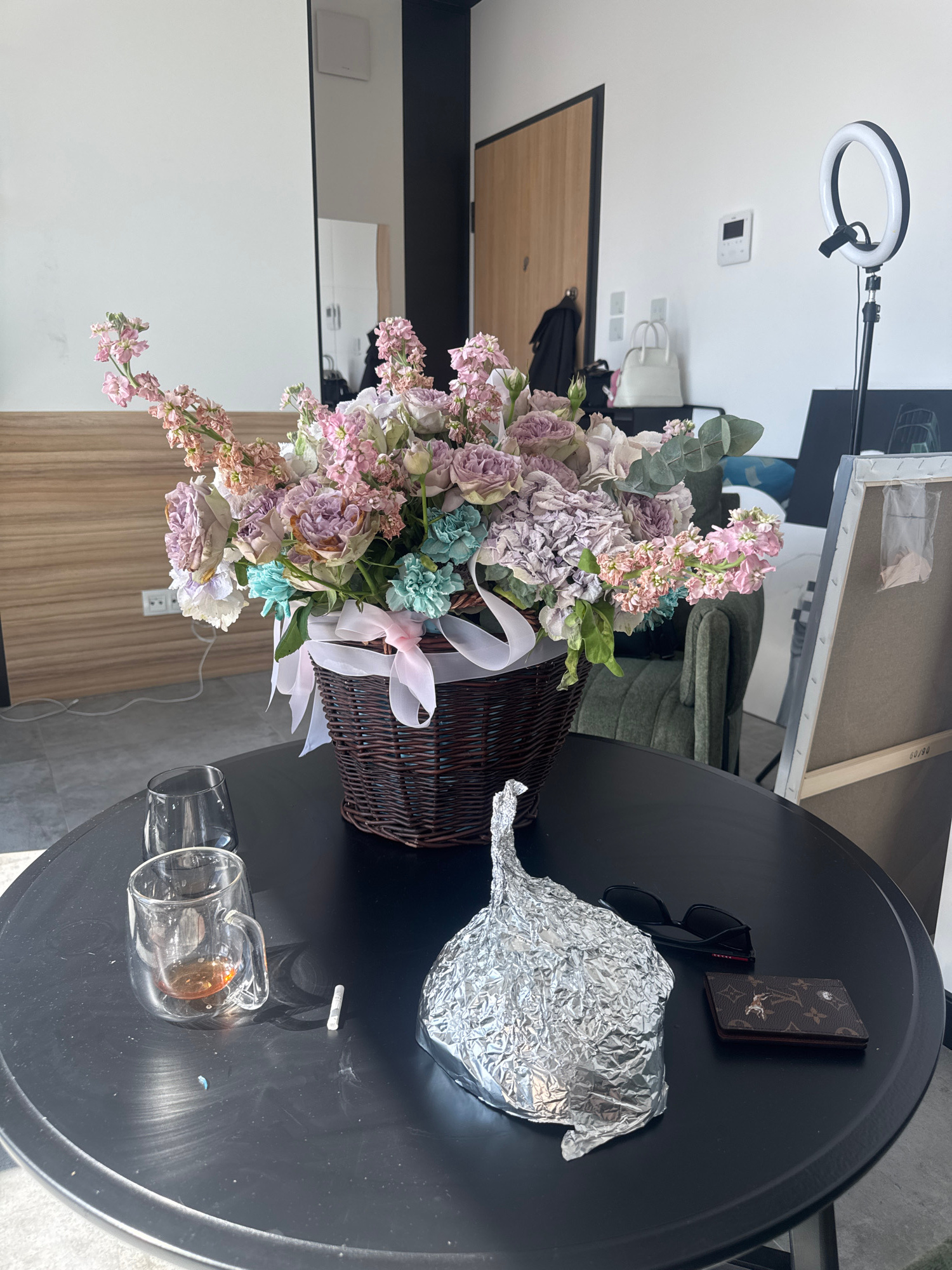 A high-angle shot shows a dark, round table with various objects on it. The primary subject is a large basket overflowing with a bouquet of flowers in shades of pink, purple, and blue. A pink and white ribbon is tied around the basket. To the right of the flowers, a crumpled ball of aluminum foil is visible. Next to the foil, a pair of black sunglasses and a small, brown wallet with a distinctive monogram pattern are placed. Further to the right, two clear glass cups, one containing a small amount of amber liquid, are present. A white object, possibly a cigarette, lies on the table between the glasses and the foil.
In the background, a ring light on a stand is positioned over a white surface. A glimpse of what appears to be a painting on an easel is also visible. The overall setting suggests an indoor space, possibly a studio or a room where a creative activity might take place. The lighting is bright, indicating it could be daytime or artificial lighting is in use. The scene is static, with no activity explicitly depicted. There is no visible text in the image. The location is specified as Warsaw, Poland.