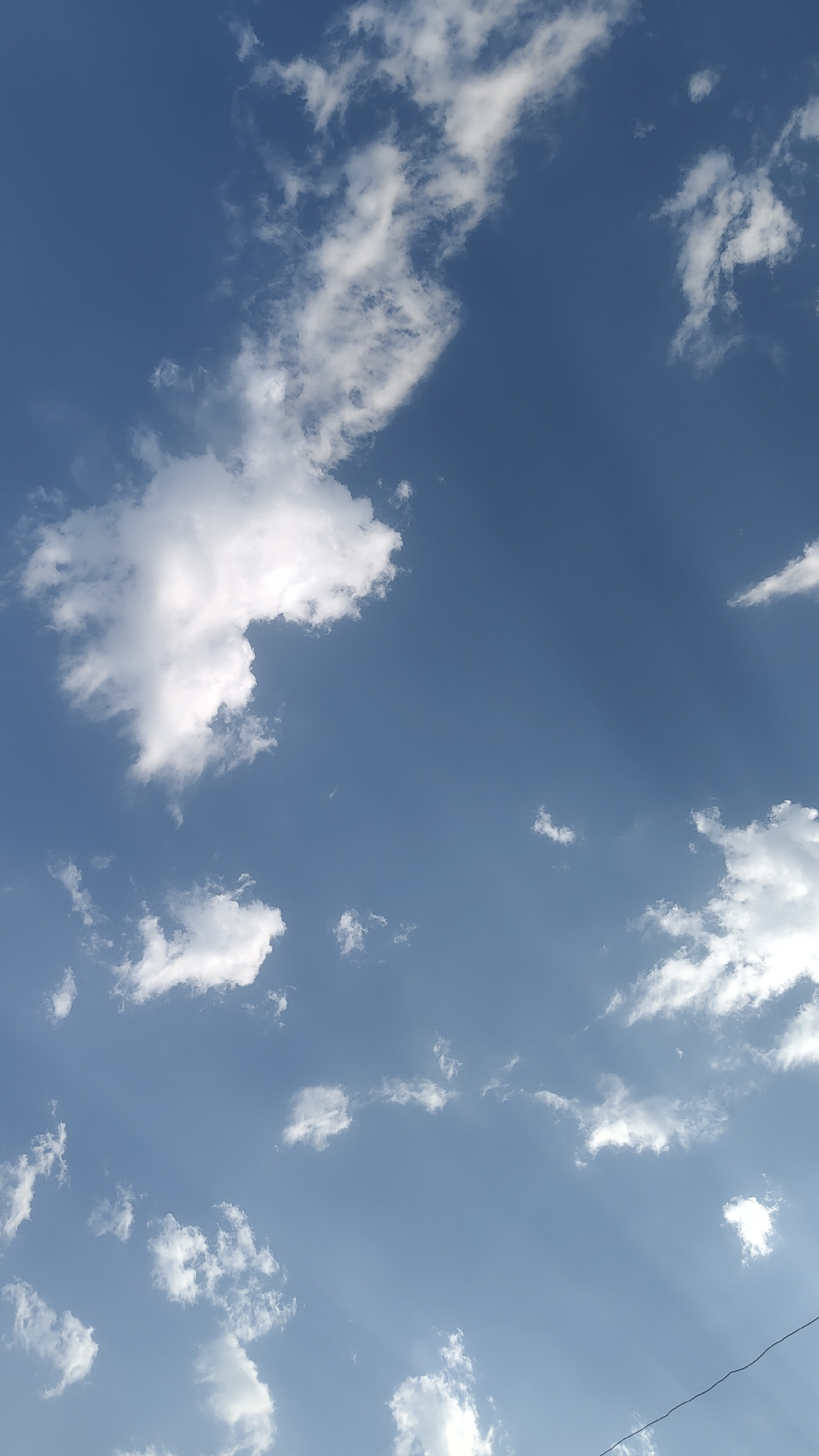 The media file captures a clear blue sky with scattered white cumulus clouds. The clouds vary in size and density, with some appearing wispy and others more defined and puffy. Sunlight is filtering through the clouds, creating subtle crepuscular rays that are visible against the blue expanse. The scene suggests a daytime setting, likely late morning or afternoon given the brightness of the sky and the quality of the light. There are no visible people or man-made structures in the majority of the frame, only a thin dark line that might be a wire or a distant object at the very bottom edge of the image. The overall impression is one of a peaceful and open sky, typical of fair weather. The location context indicates this scene is in Gulistan, Uzbekistan.