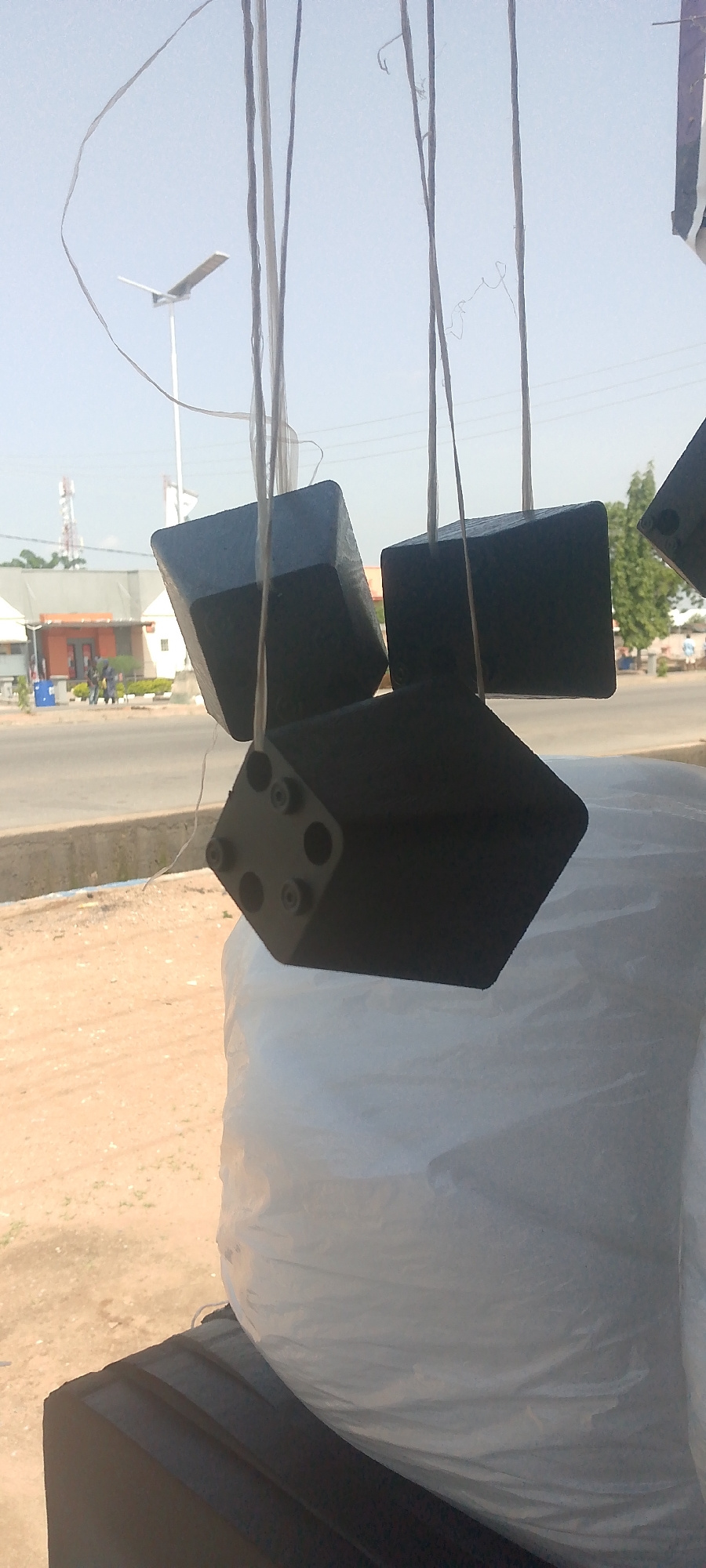 The image shows several black, angular objects, possibly speaker enclosures or sound equipment, hanging from strings against a bright sky. These objects appear to be suspended in an outdoor setting, with a street and buildings visible in the background. In the distance, a solar-powered street light is illuminated, and there are trees and communication towers. The ground in the foreground is sandy and dusty, suggesting a dry environment. A large white, plastic-wrapped object is positioned below the hanging items. The scene is likely located in Jalingo, Nigeria, given the context. There are no people clearly visible in action or interaction within the frame.