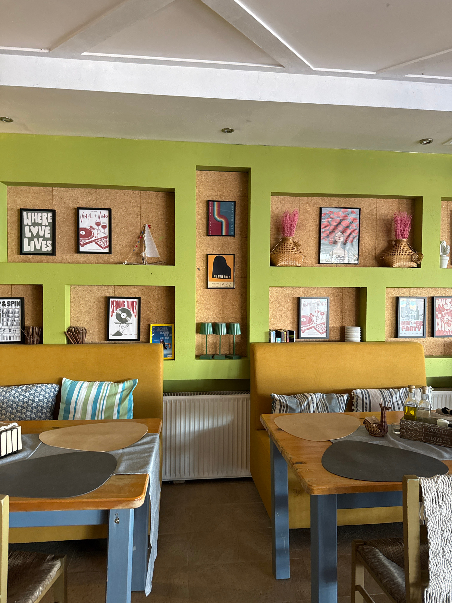 The image depicts a brightly lit, vertically oriented interior scene. The walls are painted a vibrant lime green and are adorned with framed artwork, woven baskets with pink decorative elements, and what appear to be record covers. One frame displays text that reads "WHERE LOVE LIVES." Another features a graphic of a record player and reads "VINYL + VINO." A small toy sailboat hangs on the wall.
Adjacent to the decorated wall is a mustard yellow upholstered bench or booth. On the bench are several throw pillows, one with blue and green stripes and another with a blue and white patterned design. To the right of the bench, a wooden table is visible, set with placemats. One placemat is round and light brown, while another is oval and dark gray. A small woven object, possibly a basket, sits on the table. There are also several bottles on a shelf above the table, with some text visible that includes "FAMILY" and "LOVE."
The overall impression is a casual, perhaps a cafe or restaurant setting, with a decorative and somewhat eclectic style. The lighting appears to be artificial, suggesting it could be daytime or evening. There are no people visible in the image.
