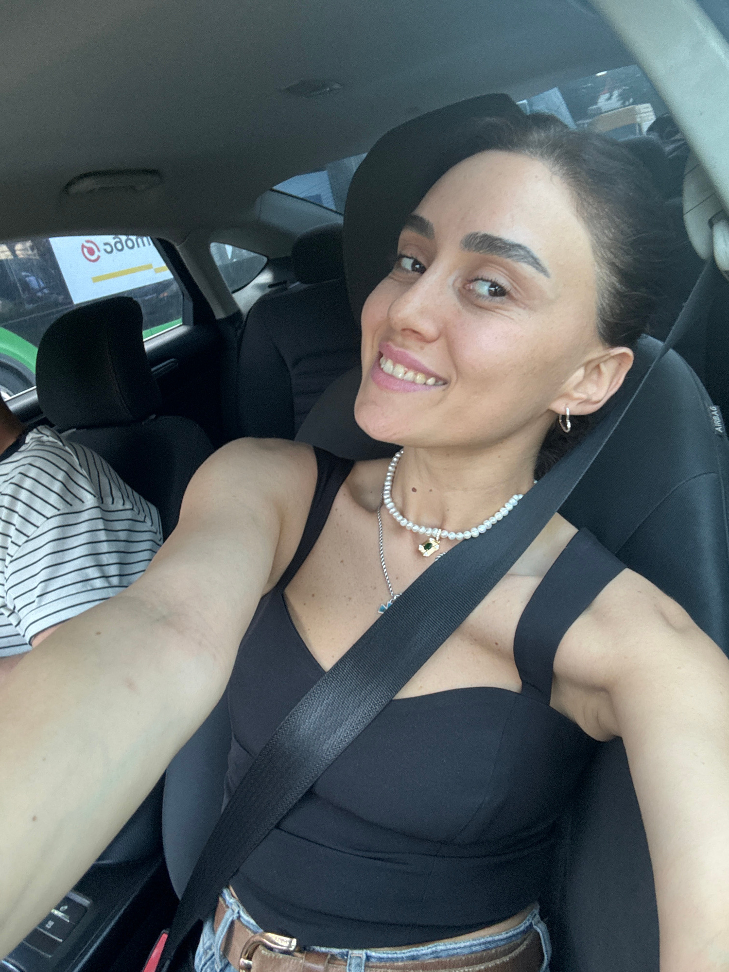 A woman with dark hair pulled back and visible eyebrows is smiling while looking towards the camera from the passenger seat of a car. She is wearing a black top, a pearl necklace with a pendant, and hoop earrings. A seatbelt is visible across her chest. To her left, a person wearing a striped shirt is partially visible. The interior of the car includes black seats and a seatbelt. In the background, through the car window, a sign with red and yellow lettering is visible, possibly indicating a business or advertisement. The car appears to be in motion in a city environment, with reflections of buildings or trees in the windows.