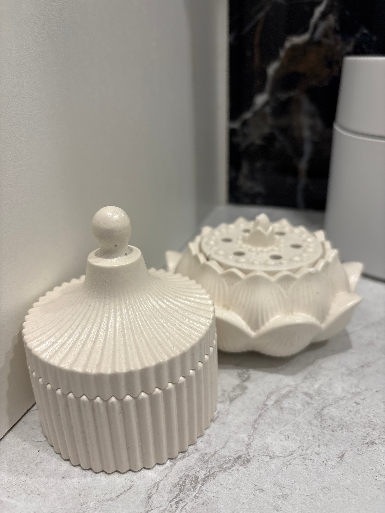 Two cream-colored decorative objects are positioned on a marble-patterned surface. The object in the foreground on the left is a lidded container with vertical fluting and a round knob on top of the lid. The object on the right is shaped like a lotus flower, with a detailed lid featuring multiple small holes. In the background, a dark, marbled surface is visible, along with a portion of a white cylindrical object.