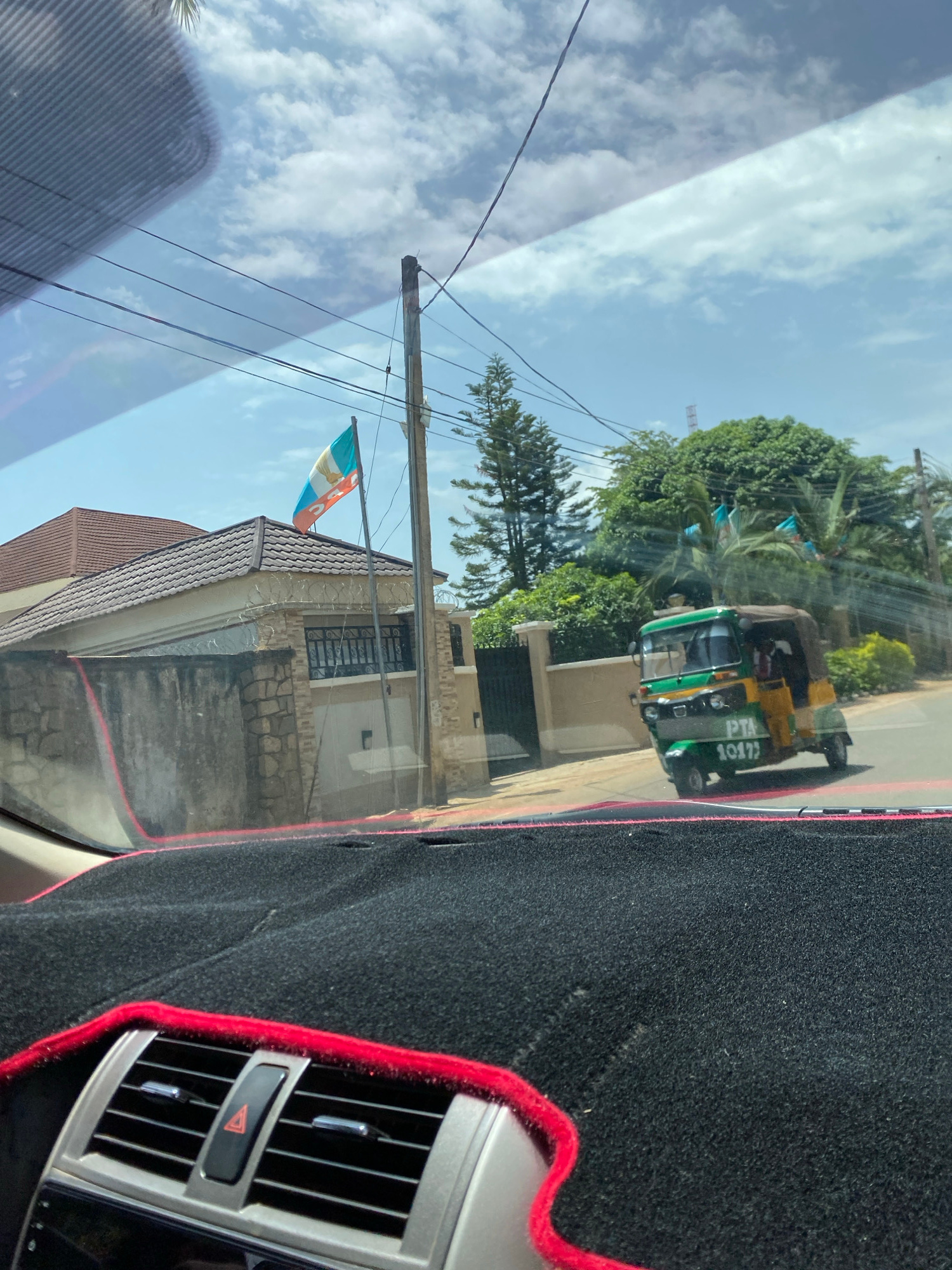 The image captures a scene through a car windshield, with a dashboard partially visible on the right. The primary subject is a green and yellow motorized rickshaw, also known as an auto-rickshaw or tuk-tuk, with visible text on its side reading "PTA 10177". The rickshaw appears to be in motion on a paved road, surrounded by lush green trees and vegetation. A building with a tiled roof and barred windows is visible behind the rickshaw, along with a utility pole and overhead wires. A flag, predominantly white with blue and orange sections and an emblem, is attached to the utility pole, fluttering in the breeze. The sky is partly cloudy with a bright, clear blue expanse. The overall lighting suggests it is daytime, likely mid-morning or afternoon, under fair weather conditions. The image seems to be taken from inside a vehicle, indicated by the reflection on the windshield and the car's interior elements. The scene suggests a typical street view in a tropical or subtropical region, potentially a town or city. The presence of the rickshaw and the style of the building hint at a location in Nigeria, as specified by the location context.