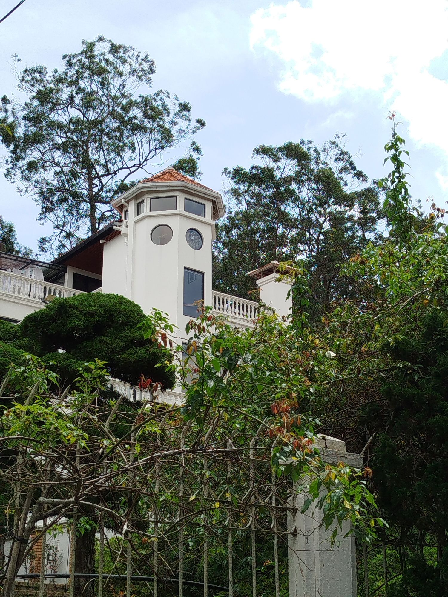 The image shows a white, octagonal tower with a terracotta roof, situated on a hillside in Da Lat, Vietnam. The tower is part of a larger building with white railings and a red-tiled roof visible to the left. Lush green trees and vegetation surround the structure, including a large tree with thin branches in the foreground and a dense green bush in front of the tower. The sky is cloudy and light blue. A metal fence with vertical bars is also visible in the lower part of the frame. The overall impression is of a residential building nestled within a natural, possibly elevated, landscape characteristic of Da Lat.