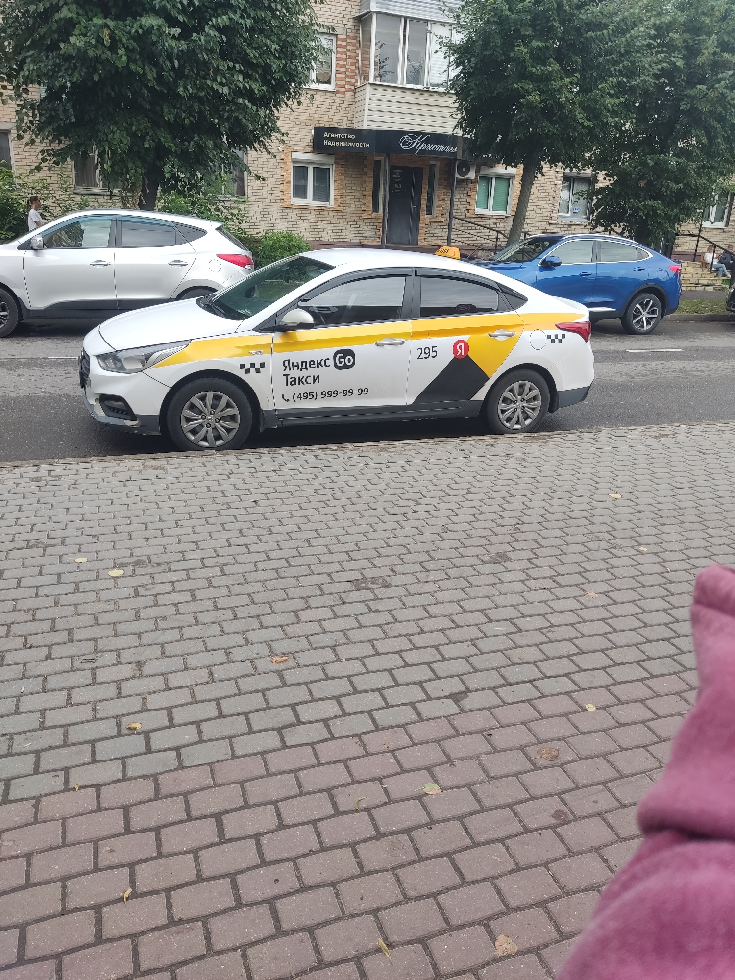 A white Yandex Go taxi is parked on the side of a street in Serpukhov, Russia, next to a brick building with several windows and trees. The taxi has a yellow stripe on its side and the Yandex Go logo. The number "295" is visible on the car door, along with a phone number. A silver SUV is parked in front of the taxi, and a blue car is parked behind it. A person can be seen standing near the silver SUV. The street is paved with cobblestones.