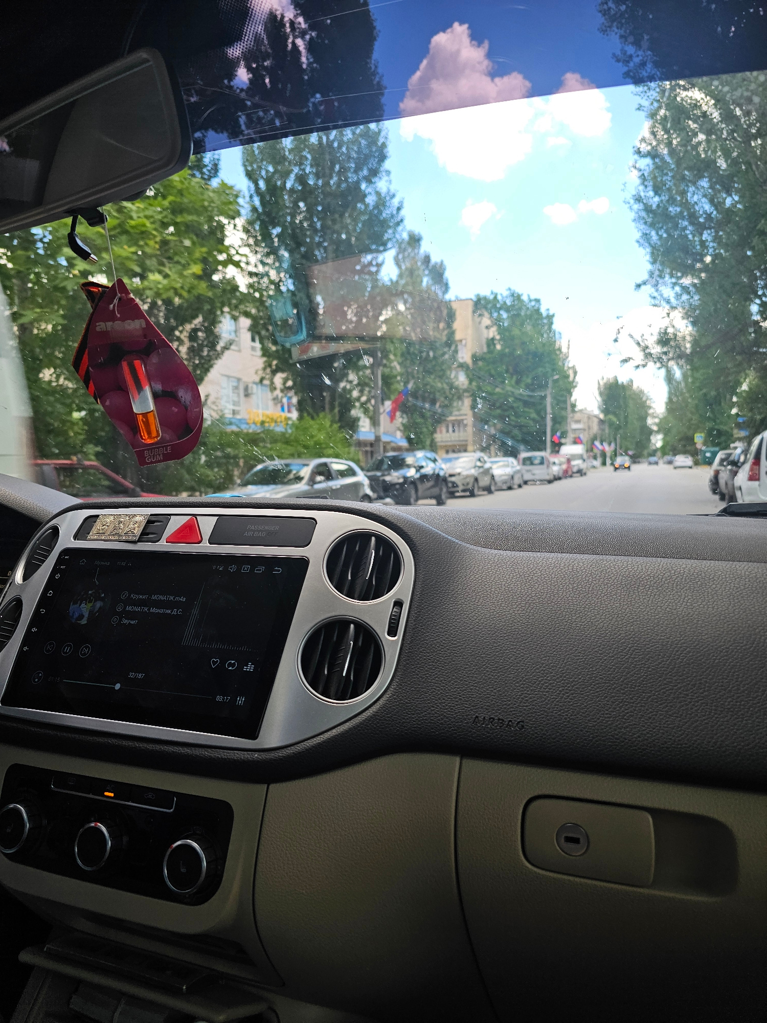 The image is taken from inside a vehicle, looking out through the windshield. A car air freshener shaped like lips with a red and black pattern hangs from the rearview mirror. The dashboard is visible, with the car's infotainment screen displaying song information and playback controls. To the right of the screen are air vents. Below the screen are climate control knobs. The car is on a street lined with trees and parked cars. The sky is blue with white clouds. The scene is consistent with a street in Skadovsk, Ukraine, as indicated by context.