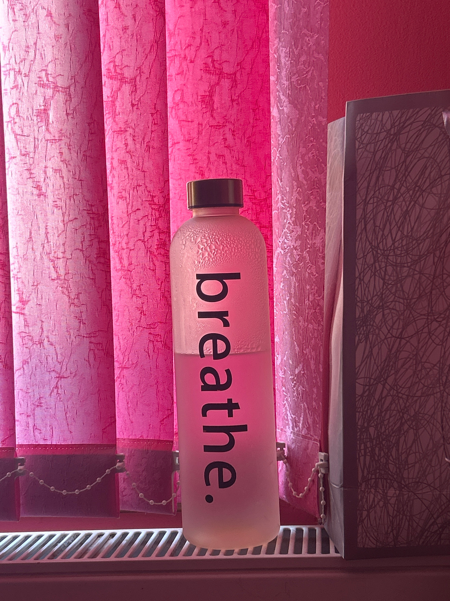 A frosted glass water bottle with the word "breathe." printed vertically in black text stands on a white radiator. The bottle is half-filled with a light pink liquid and condensation is visible on the upper portion of the bottle. The background consists of pink vertical blinds with a textured pattern and a dark rectangular object to the right of the bottle. The image appears to have been taken indoors. The location is Knezha, Bulgaria.