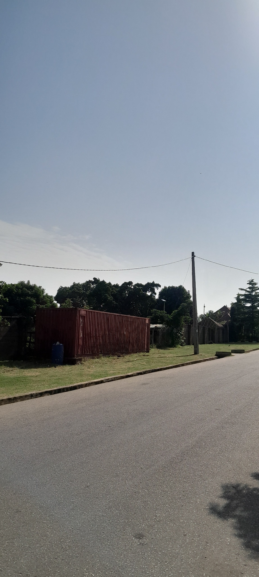 A static, eye-level, vertical, full shot captures a desolate roadside scene under a clear, pale blue sky. In the foreground, a gray asphalt road curves gently to the right, casting a dappled shadow of tree foliage on its surface. Along the left edge of the road, a concrete curb separates it from a strip of grass. To the left of the grass, a large, rusted, red corrugated metal shipping container is positioned, with a blue barrel standing beside it. Beyond the container, a dense line of dark green trees forms a backdrop. To the right of the trees, a tall, weathered wooden utility pole stands, with electrical wires extending horizontally across the frame. Further to the right, glimpses of dilapidated structures with corrugated metal roofs are visible, partially obscured by the trees and foliage. The overall atmosphere is one of neglect and emptiness, typical of a peripheral area within the city of Jalingo, Nigeria, on a bright, cloudless day.
