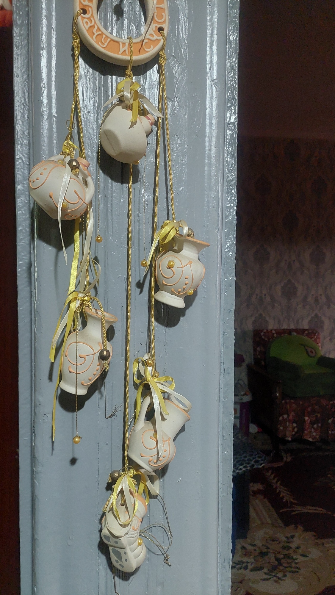 The image shows a decorative ceramic hanging ornament with multiple small, white, jug-shaped pieces adorned with orange swirls and yellow ribbons, suspended from a larger horseshoe-shaped ceramic piece with orange lettering. The ornament is hanging on a gray painted wooden surface, likely a door or frame. In the background, out of focus, is a room with patterned wallpaper and furniture, including a dark chair with a green cushion and a rug. The scene is lit from the side, casting shadows. This image was taken in Angren, Uzbekistan.