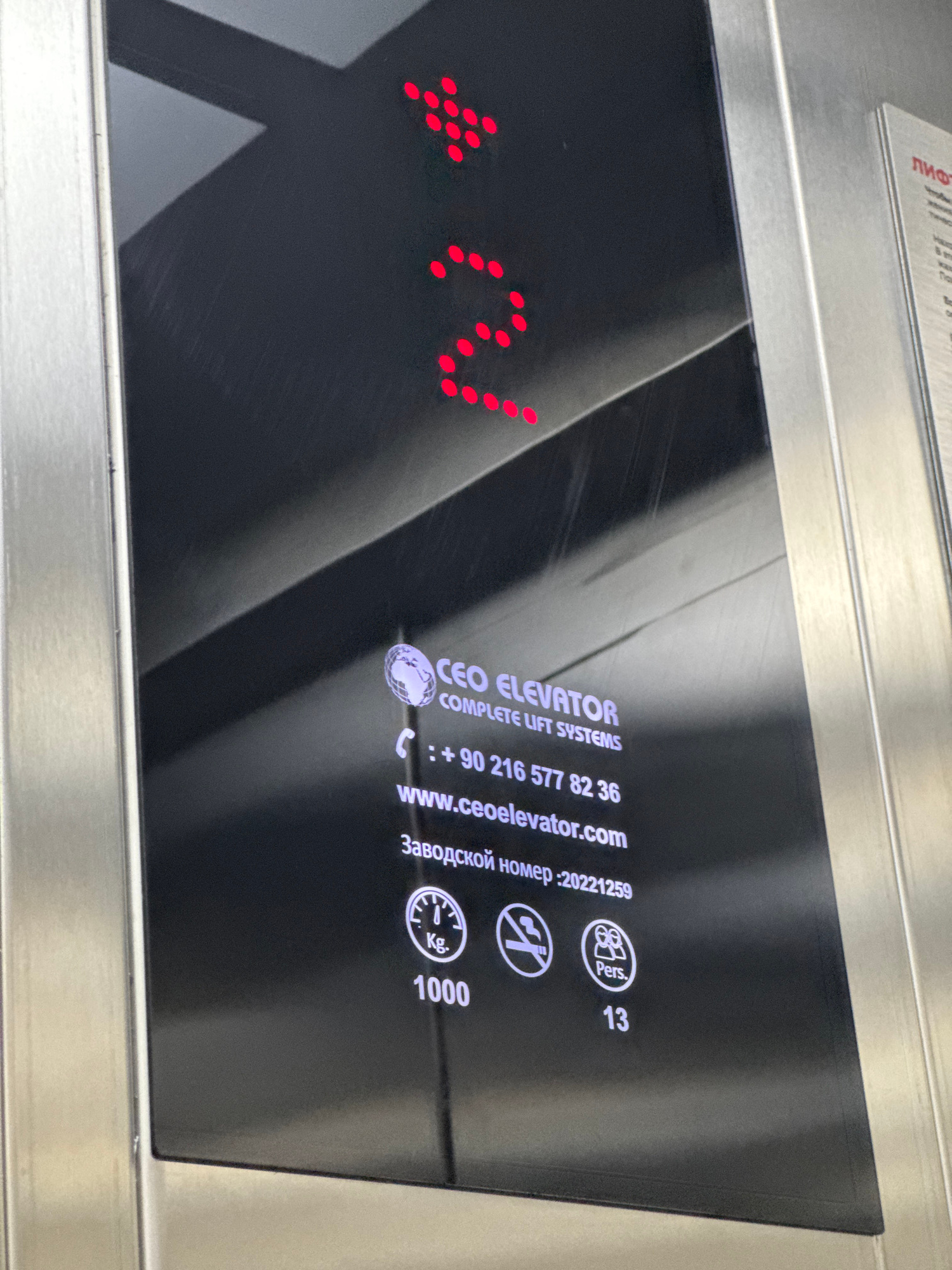 The image shows an elevator control panel. The floor indicator displays the number "2" in red LEDs. Below the floor indicator, there is a logo for "CEO ELEVATOR COMPLETE LIFT SYSTEMS", a phone number "+90 216 577 82 36", a website "www.ceoelevator.com", and a reference number ":20221259". There are also icons indicating weight capacity (1000 Kg) and passenger capacity (13 Pers.), as well as a "no smoking" symbol. The panel is made of brushed metal and black glass. There is text in Russian below the website address. A separate information sheet with Russian text, partially visible on the right side of the image, is also present. The lighting suggests an indoor environment. The specific location in Mikhaylovsk, Russia, is not identifiable from the visual information alone.