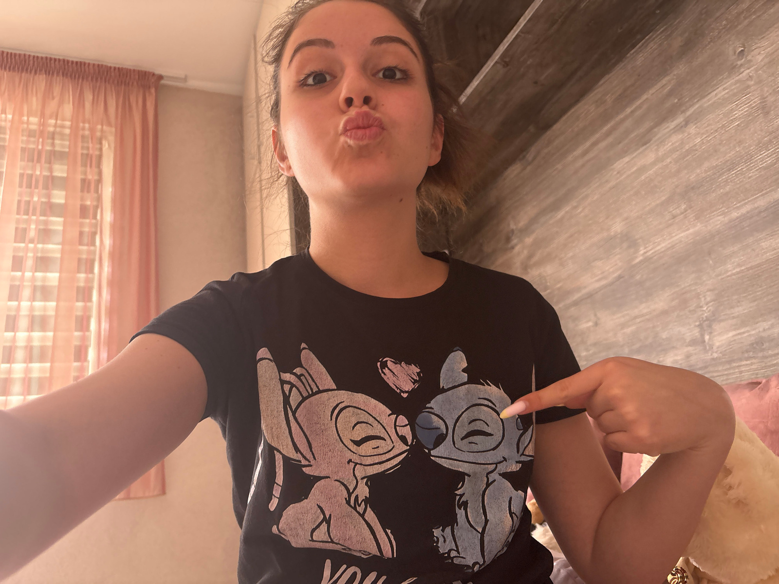The image shows a young woman with her lips puckered, making a kissing face. She is wearing a black t-shirt with a graphic of two Stitch-like characters kissing, with a pink heart above them. The woman is pointing at the graphic on her shirt with her right hand. She has long, dark hair and is looking directly at the camera. The background is blurred, but appears to be a bedroom with a pink curtain on the left and a wooden headboard on the right. The lighting is soft and warm. The image was taken indoors, likely in a residential setting. There is no information to suggest the location is in the city of Bulgarovo, Bulgaria.