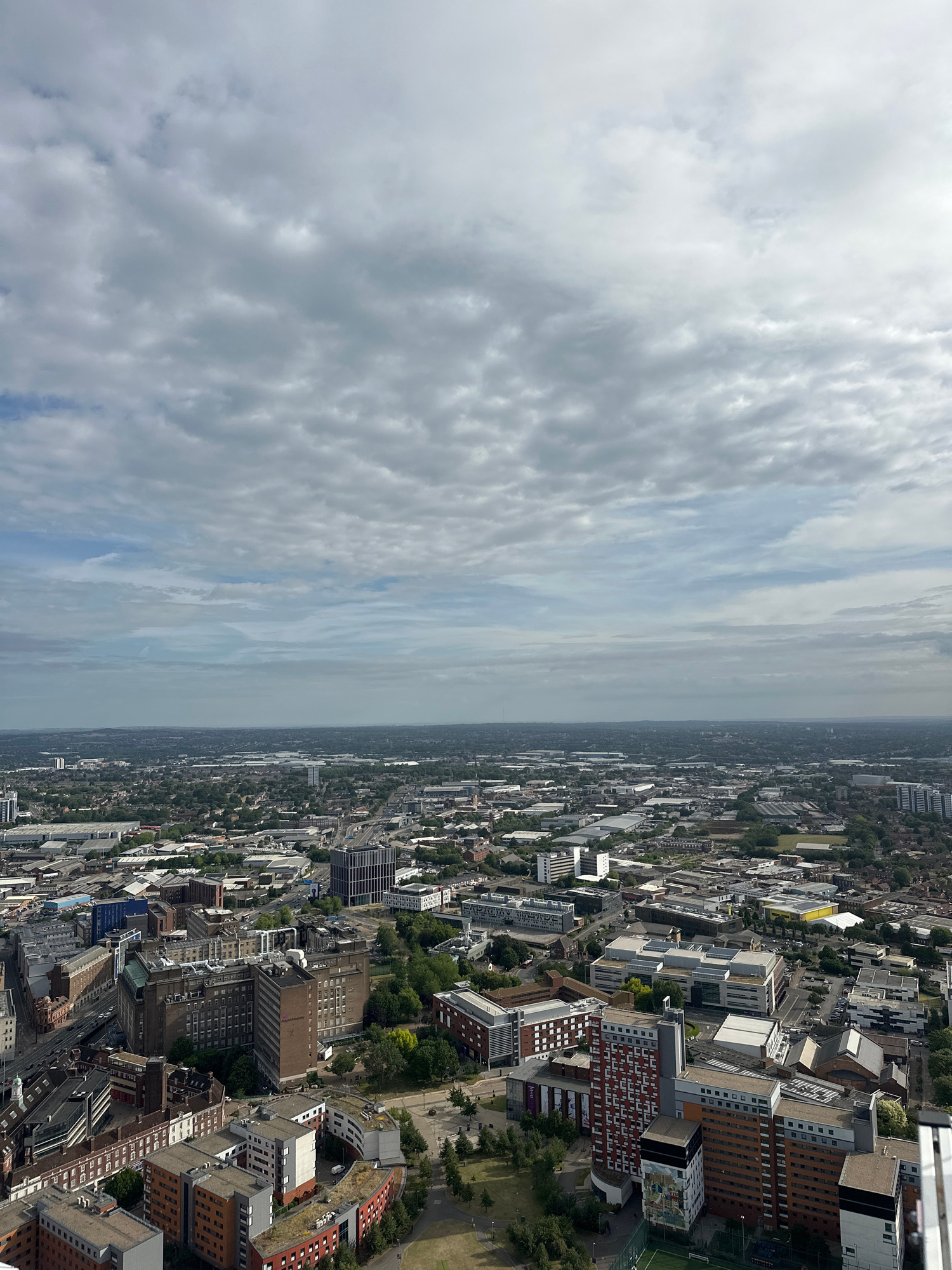 An aerial view captures a sprawling cityscape under a cloudy sky. The image showcases a dense urban environment with numerous buildings of varying heights and architectural styles, interspersed with patches of green trees and parks. Roads and infrastructure are visible throughout, indicating a well-developed city. The foreground features several distinct buildings, including multi-story residential blocks with modern designs, some featuring colorful facades, and older, larger brick structures. In the mid-ground and background, the cityscape extends towards the horizon, with industrial buildings, smaller residential areas, and larger complexes. The sky is filled with a textured layer of gray and white clouds, suggesting overcast or partly cloudy weather. This view is consistent with the urban landscape of Birmingham, United Kingdom, likely taken from a tall building or viewpoint overlooking the city center and surrounding districts. No individuals or specific actions are discernible from this elevated perspective.
