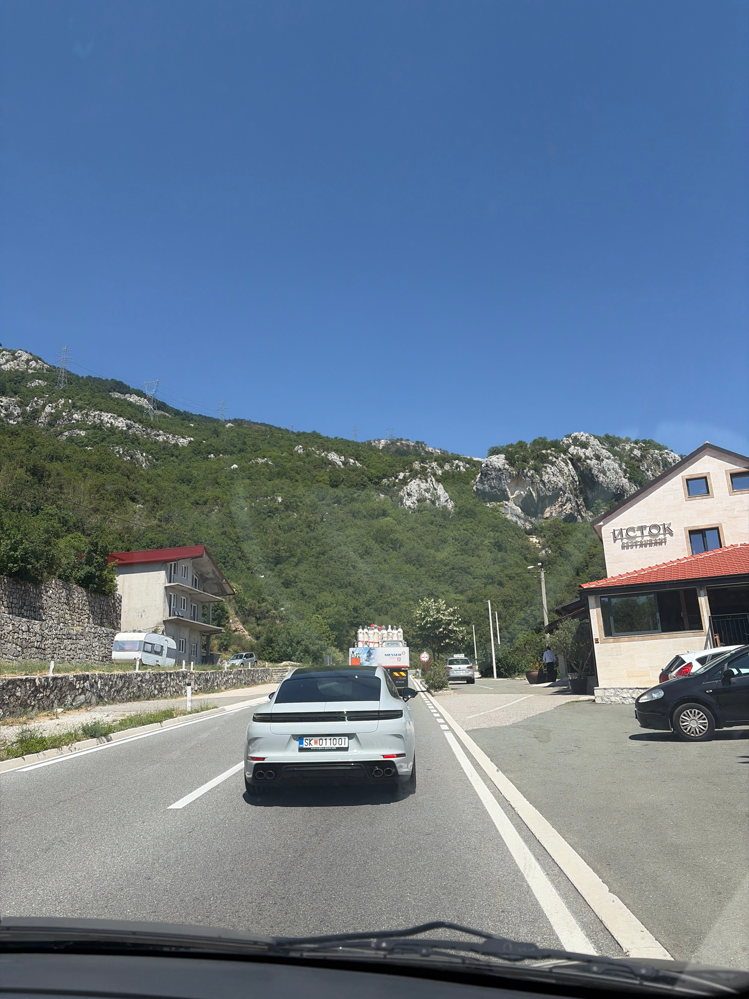 The image shows a view from a vehicle driving on a road next to a steep, forested mountainside. A white Porsche SUV is in front, driving on the same road. To the left of the road is a building with the name "ISTOK RESTORAN" visible. Further up the mountainside, more buildings and power lines can be seen. The sky is clear and blue, suggesting a sunny day. The scene appears to be in Lapčići, Montenegro. The time of day is likely midday due to the bright sunlight and clear sky. The overall impression is that of a scenic drive through a mountainous region.