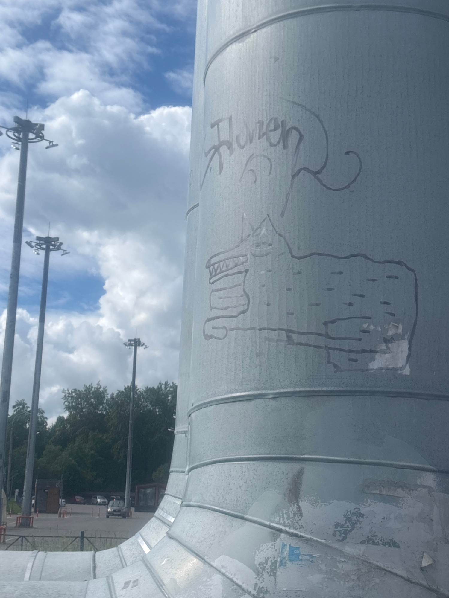 The image shows a large, cylindrical metal structure with graffiti on its surface. The graffiti includes text that appears to read "Homen" and a drawing of a creature that resembles a wolf or a dog. The structure is situated outdoors, with a cloudy sky and trees in the background. In the distance, there are tall light poles and some vehicles, suggesting an urban or semi-urban environment. The metal surface of the structure shows signs of weathering and some peeling paint or material. The overall impression is of an outdoor industrial or public space marked by graffiti.
