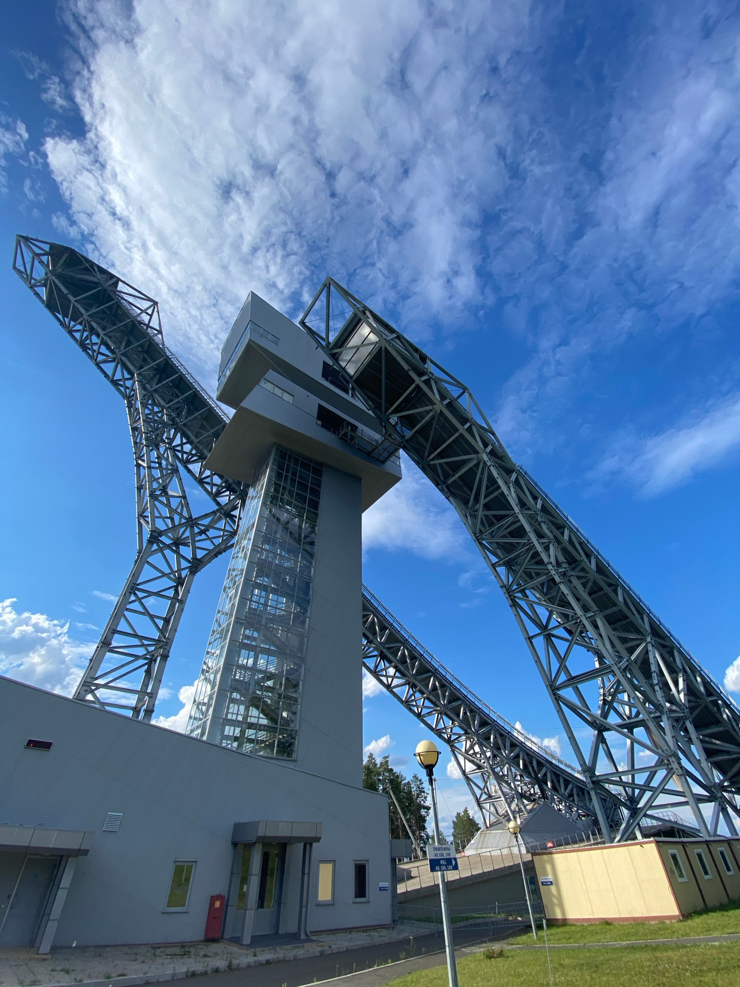 This low-angle shot captures a large, modern industrial structure against a bright blue sky filled with scattered white clouds. The structure appears to be a bridge or a large elevated pathway, constructed with intricate metal latticework that forms a complex geometric pattern. The perspective emphasizes the scale and industrial design of the object.
A central tower-like element rises from the structure, featuring a sleek, grey facade and large glass windows that reflect the sky and the metal framework. The tower has a more contemporary architectural style compared to the exposed metal trusses. Adjacent to the tower, a building with a light yellow facade and dark-trimmed windows is visible, suggesting an operational facility or complex.
The scene is likely taking place during the daytime, given the bright lighting and clear sky with clouds. The weather appears fair and sunny. A sign with Cyrillic text is visible near the base of the structure. The overall impression is one of modern engineering and infrastructure within an outdoor setting. The image evokes a sense of industrial grandeur and the vastness of the sky.