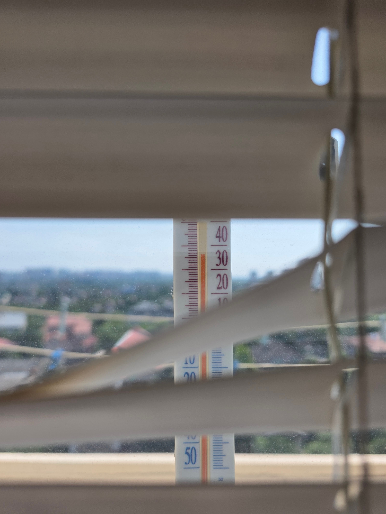 This media file is a close-up photograph of a thermometer positioned behind partially drawn vertical blinds, suggesting an indoor setting looking outwards. The thermometer displays temperature readings in Celsius, with visible markings for 20, 30, 40, and 50 degrees. The mercury level on the left thermometer is between 30 and 40 degrees, closer to 30. The thermometer on the right shows a reading around 50 degrees. The background, seen through the blinds and the thermometer, depicts an outdoor scene with greenery, suggesting trees and possibly residential buildings with reddish rooftops, indicative of a residential area. The sky appears light and hazy, hinting at daylight, possibly a warm or hot day given the high temperature readings on the thermometer. The overall impression is that of a domestic environment in Kyiv, Ukraine, with the thermometer providing a specific data point about the ambient temperature. There are no people visible, and the focus is on the thermometer and its readings within the context of the window view. The image is slightly blurry, particularly the background, drawing attention to the thermometer itself.