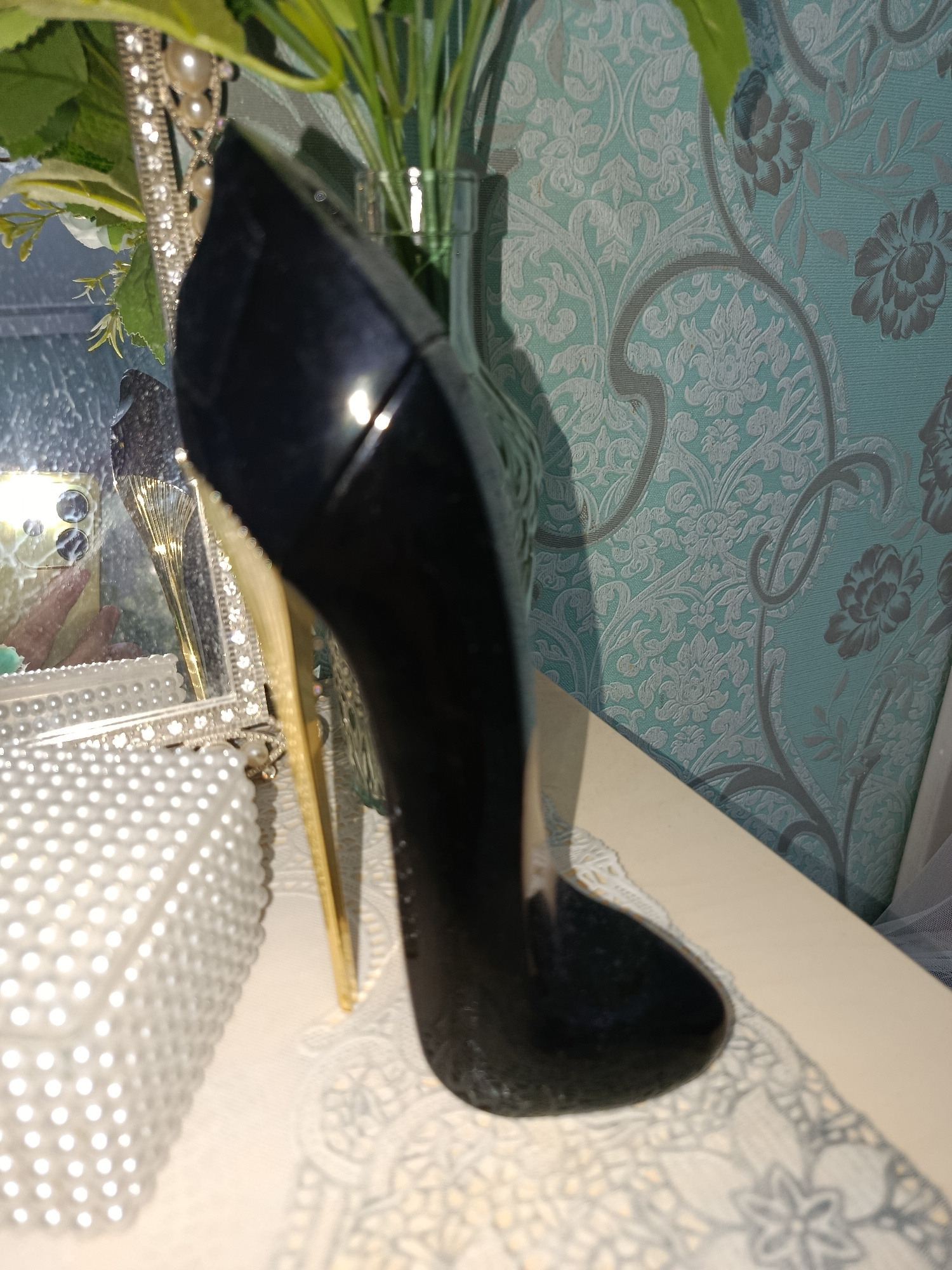 A black high-heeled shoe is positioned on a light-colored surface, casting a shadow. In the background, a patterned wallpaper in shades of teal and grey is visible, along with a vase containing green foliage. To the left of the shoe, a decorative box covered in white beads and a mirrored frame with ornate detailing are present. The lighting in the image creates reflections on the shoe and other surfaces. The scene appears to be an indoor setting, possibly a room. There are no people or actions depicted. The location within the city of Tashkent, Uzbekistan cannot be determined from the provided image.