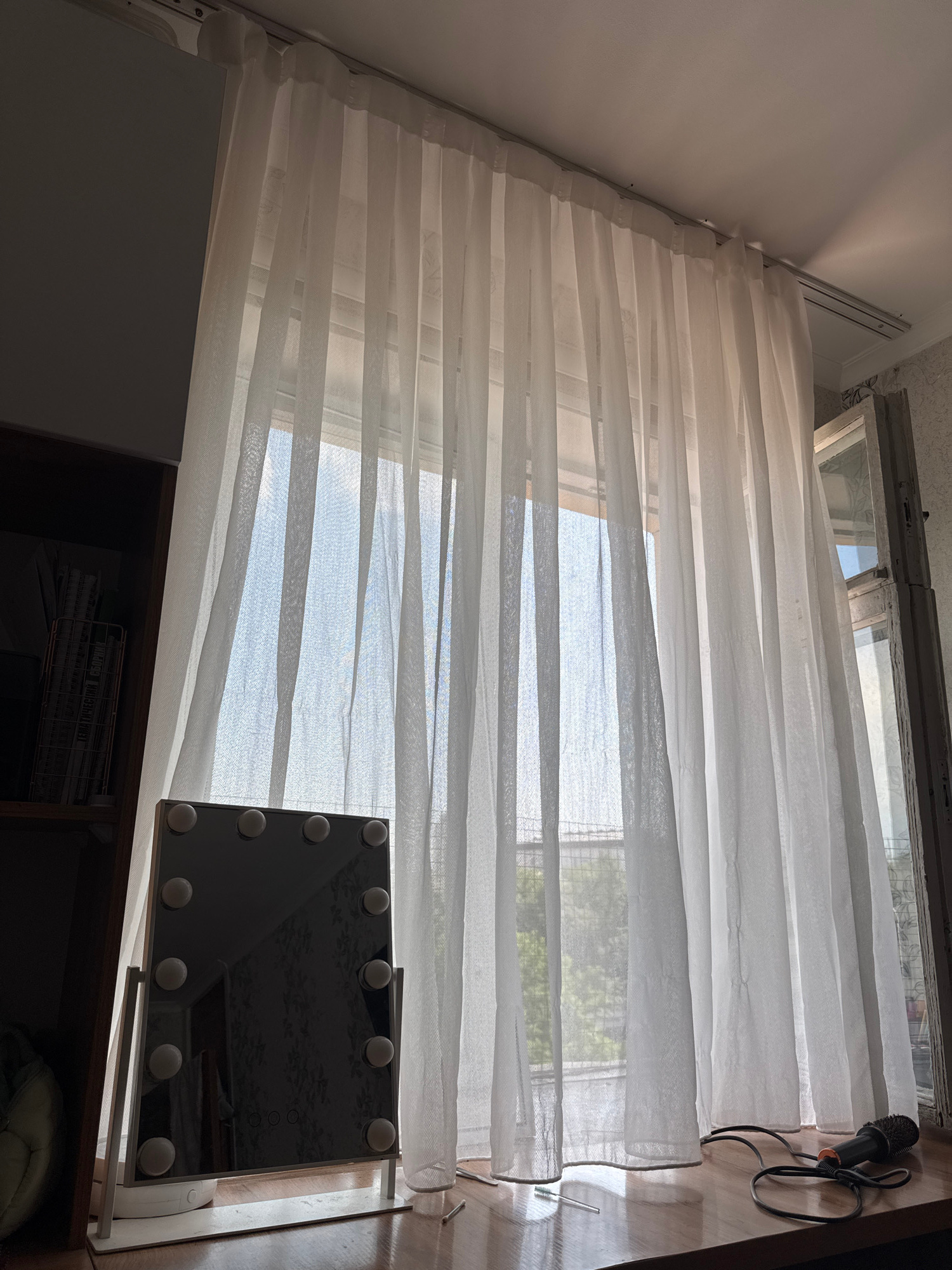 An indoor scene depicts a window covered by sheer white curtains, allowing diffused sunlight to enter. On a wooden surface in front of the curtains, a lighted makeup mirror with multiple bulbs stands on its base. To the right, a black microphone with a coiled cable rests on the same surface. To the left, a dark wooden shelf unit is partially visible, filled with books and other items. The overall environment suggests a domestic setting, possibly a bedroom or vanity area.