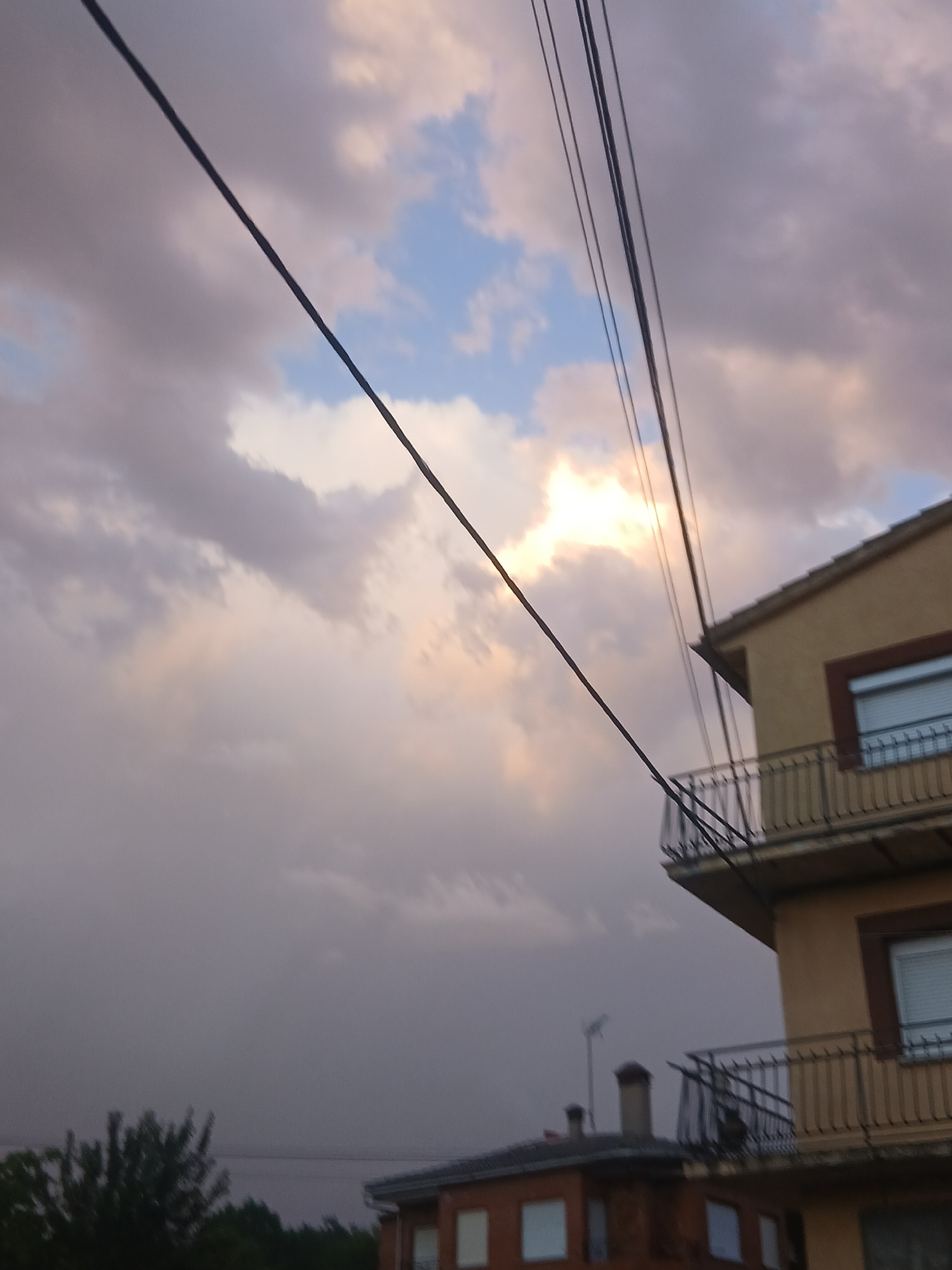 The image shows a cloudy sky with some sunlight breaking through. There are power lines crossing the sky diagonally. In the foreground, the corner of a building with balconies is visible. Another building is partially visible to the left, with trees in front of it. The scene appears to be an outdoor urban environment, possibly Navaluenga, Spain, given the architectural style.