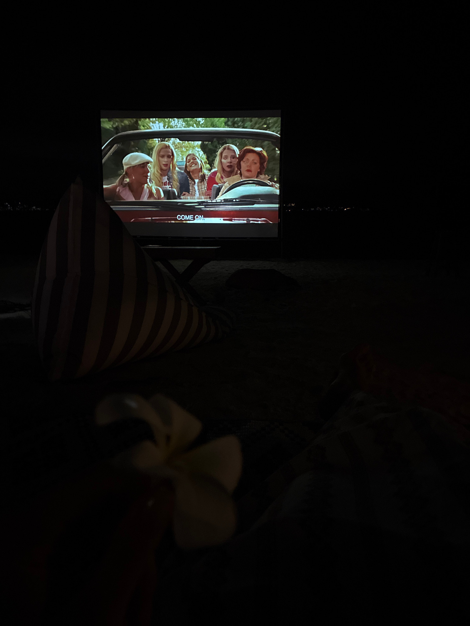 A night scene shows an outdoor movie projection on a large screen, with a beanbag chair in the foreground. The movie displays a group of four young women in a car, with the text "COME ON" visible on the screen. In the lower foreground, a white flower is slightly out of focus on a dark surface, possibly sand. In the background, distant lights suggest a populated area. The scene is set in the Ko Pha-ngan District, Thailand.