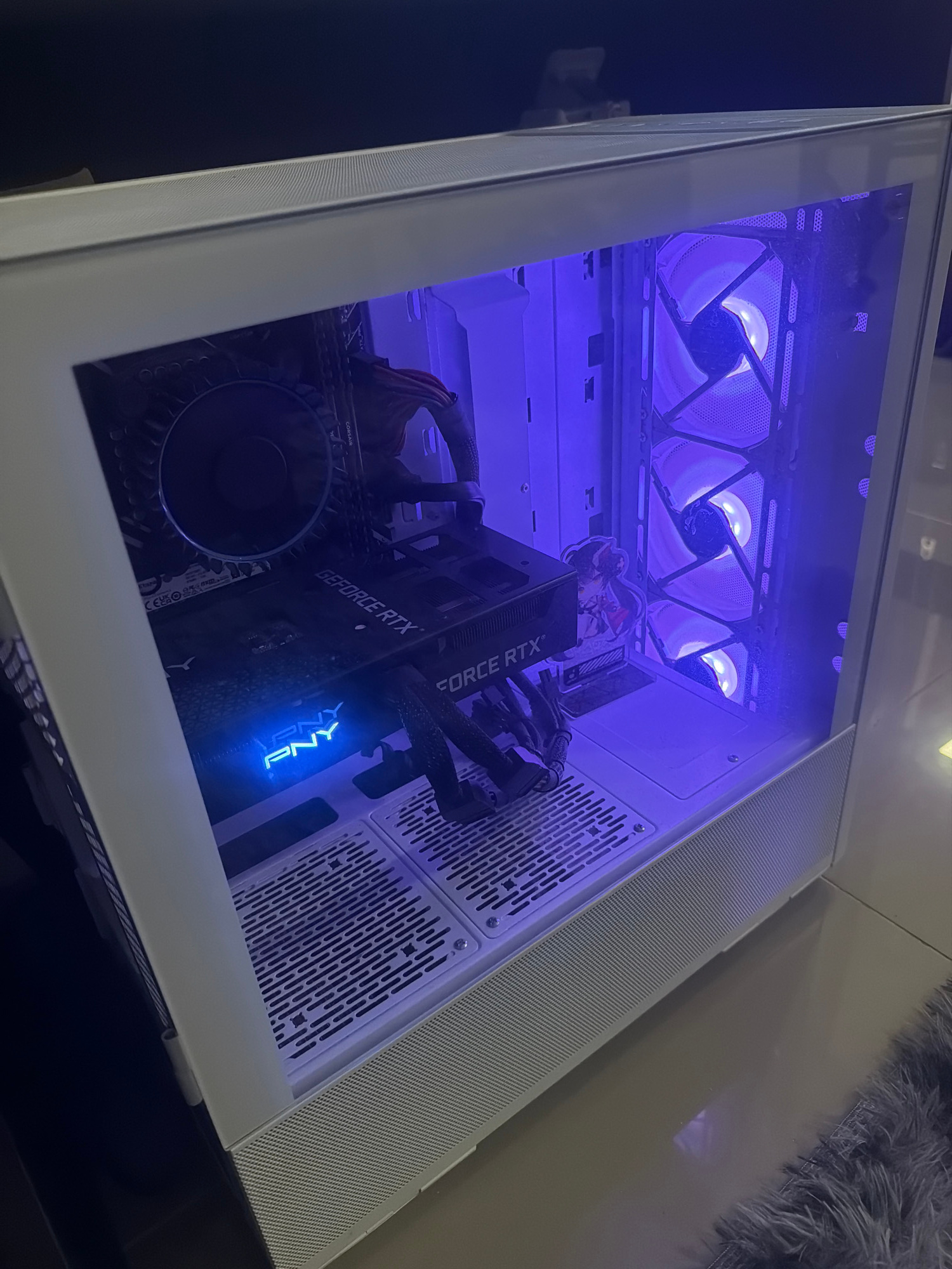 A white computer tower with a clear side panel sits on a tiled floor next to a dark, plush rug. The inside of the computer is illuminated by purple and blue LED lights. Three large fans are visible at the top of the tower, emitting a purple glow. A graphics card, labeled "GEFORCE RTX" and "PNY," is mounted vertically, with a blue PNY logo visible. The computer tower is angled towards the right side of the frame.