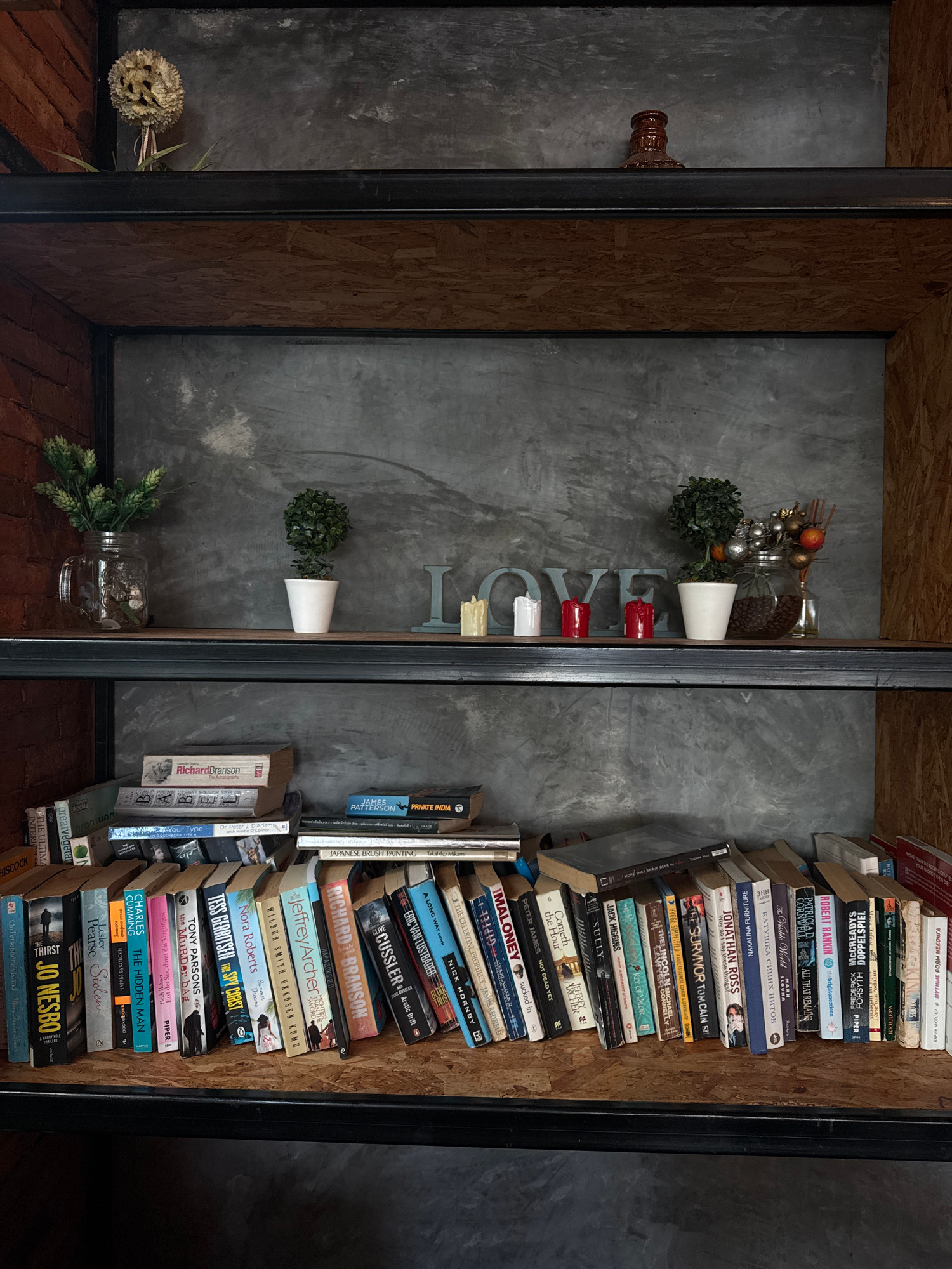 The image depicts a two-tiered shelf unit in what appears to be a cafe or shop in Rawai, Thailand. The shelves are made of dark metal and wood-like composite material, with a concrete-textured backdrop.
The top shelf displays decorative items: a dried flower arrangement on the left, a small artificial plant in a white pot in the center, and to the right, a jar filled with coffee beans, another artificial plant, and decorative baubles. The word "LOVE" is formed by light blue metal letters on the backdrop, flanked by three candles.
The bottom shelf is filled with a collection of books. They are arranged in a slightly haphazard manner, some standing upright, others leaning. Visible book titles include "Richard Branson: The Autobiography," "JAMES PATTERSON: PRIVATE INDIA," "JAPANESE BRUSH PAINTING," "THE THRST," "JO NESBO," "STOLEN," "THE HIDDEN MAN," "THE SPY CHIES," "THE SURVIVOR," and "ALL THAT REMAINS." The variety of titles suggests a personal or curated collection rather than a commercial bookstore.
The overall aesthetic is rustic and modern, with exposed brickwork visible on the far left, hinting at the building's construction. The lighting appears to be artificial, casting a warm glow on the objects. There are no people visible in the image.