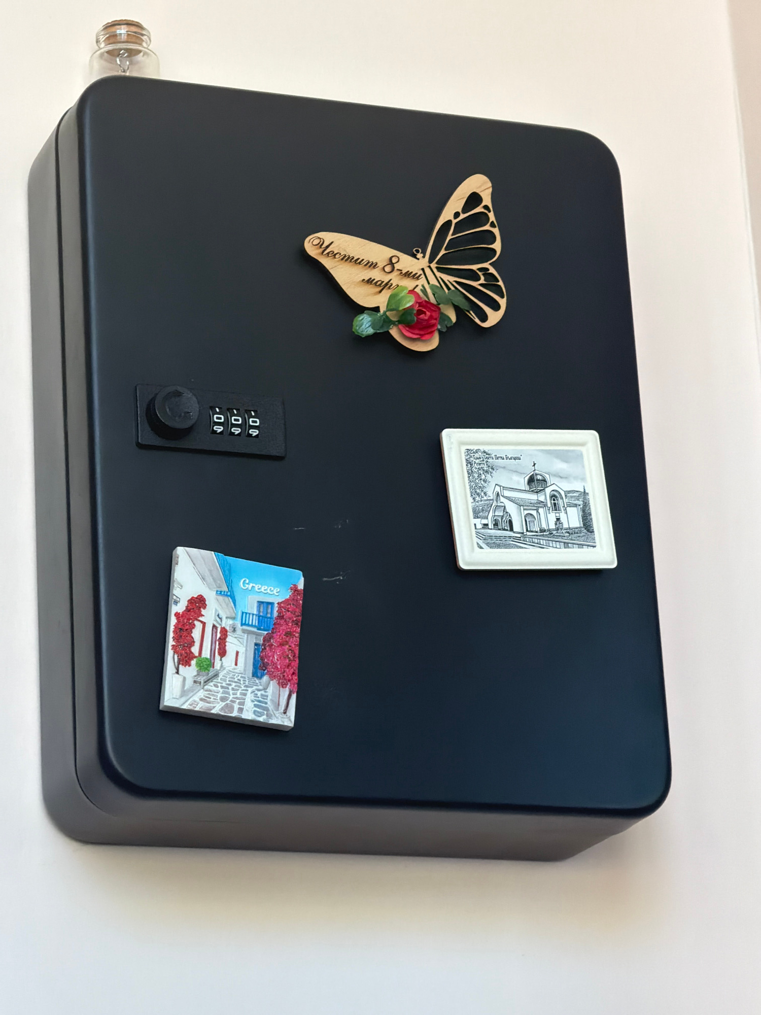 The image shows a black, rectangular lockbox mounted on a white wall. On the top of the lockbox is a small glass bottle with a cork stopper. Attached to the lockbox are three decorative items: a wooden butterfly with flowers and text, a small square magnet depicting a street scene in Greece with the word "Greece" printed on it, and a square magnet featuring a black and white drawing of a church, likely in Bulgaria, with text above it. The lockbox also has a combination lock. The scene is brightly lit, and the background is a plain white wall. There is no indication of the city of Dolno Ablanovo, Bulgaria.