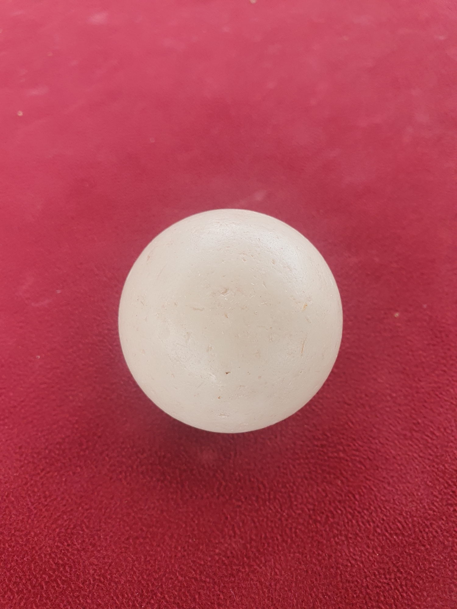 A single, off-white, spherical object is centered on a textured, deep pink surface. The object appears to be a bead or a small ball, with a slightly matte finish and some subtle imperfections or markings visible on its surface. The pink surface beneath it has a subtle, woven texture, giving it a fabric-like appearance. The lighting in the image is soft, creating a gentle shadow that is not clearly defined due to the angle. The scene is simple, focusing entirely on the interaction between the spherical object and the textured background. There are no people, animals, or specific environmental cues to suggest a particular time of day, weather, or location beyond what can be inferred from the general presentation. The image is a close-up, top-down view of these two elements. There is no visible text.