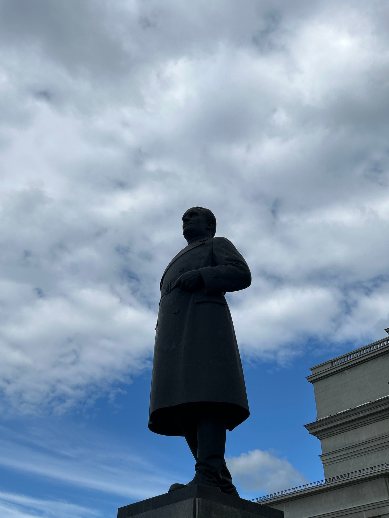 A low-angle shot captures a dark statue of a man in a long coat standing against a cloudy blue sky. The statue is positioned on a dark pedestal, and its left side is angled towards the viewer. To the right of the statue, a portion of a large, grey, multi-story building with visible architectural details and a railing is visible. The sky is filled with a mix of white and grey clouds, with patches of blue sky peeking through. The image appears to be taken outdoors in a public space. This is a statue of Ivan Ivanovich Nesterov, located in Novokuybyshevsk, Russia.