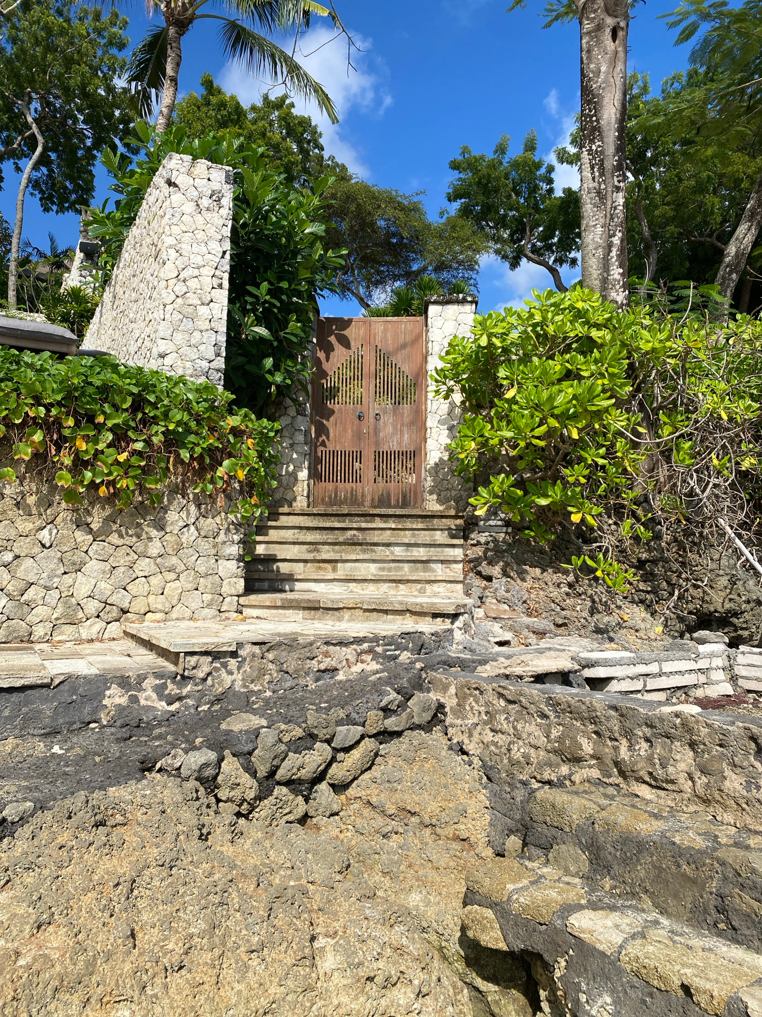 This image depicts a weathered wooden double gate, set within a textured stone wall, leading up a series of stone steps. Lush green foliage spills over the top of the wall to the left and frames the gate on the right, with trees and a palm tree visible in the background under a bright blue sky with scattered white clouds. The foreground is dominated by rough, rocky terrain and what appears to be eroded stone structures. The scene suggests a tropical, possibly coastal, environment. This location is identified as being in Kuta Selatan, Indonesia.