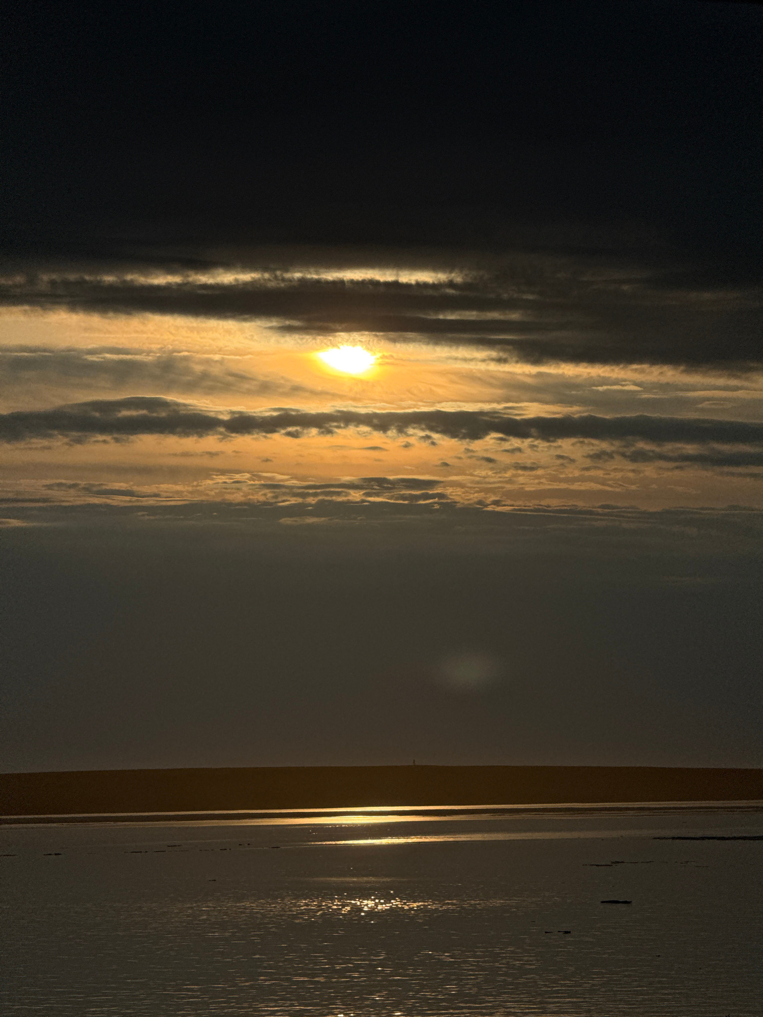 This image captures a sunset or sunrise over the sea, viewed through a window or doorway. The sun, a bright orb, is partially obscured by clouds, casting a warm, golden light across the sky. The clouds are streaked with darker shades, creating a dramatic contrast with the illuminated portions. The sea is visible in the background, appearing as a dark expanse beneath the colorful sky.
On the right side of the image, a window frame or door jamb is visible, with vertical lines suggesting a pane of glass or a textured surface. The light reflects off this surface, creating a shimmering effect. The overall mood is serene and contemplative, with the natural beauty of the sky dominating the scene. The time of day is clearly either dawn or dusk, given the low angle of the sun and the quality of light. The location is suggested to be Pevek, Russia, a coastal city, which aligns with the presence of the sea. There is no visible text in the image.