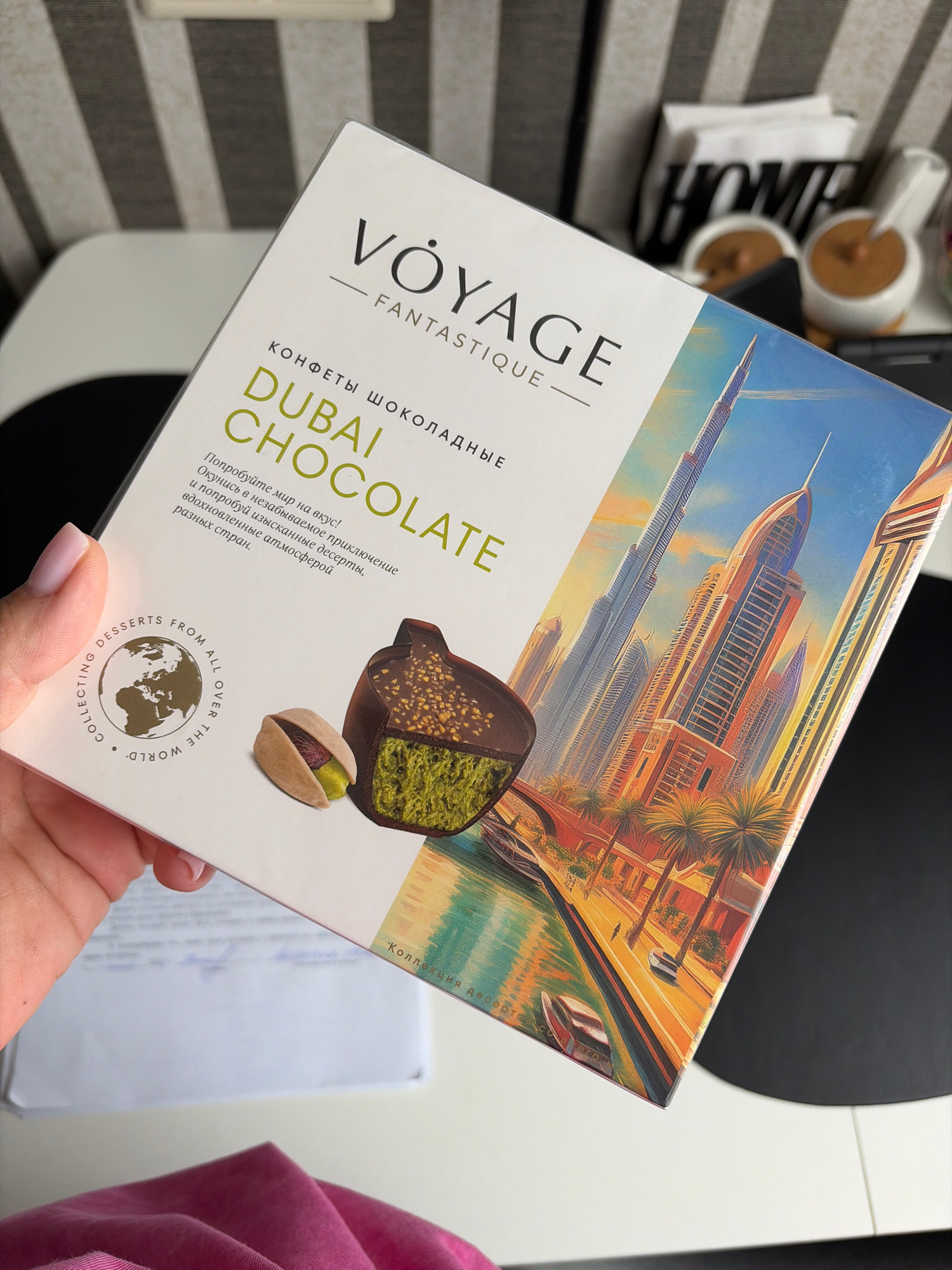 The image shows a box of "VÔYAGE FANTASTIQUE" chocolates with a "DUBAI CHOCOLATE" theme. A hand with light pink nail polish is holding the box. The box features an illustration of Dubai's skyline with skyscrapers and palm trees, a canal with boats, and a close-up of a chocolate with pistachio filling and a whole pistachio. Text on the box reads "КОНФЕТЫ ШОКОЛАДНЫЕ DUBAI CHOCOLATE" and "Попробуйте мир на вкус! Окунитесь в незабываемое приключение и попробуйте изысканные десерты, вдохновленные атмосферой разных стран." and "COLLECTING DESSERTS FROM ALL OVER THE WORLD." There is also a partial view of a white envelope with some blue writing on it. The background is blurred, showing striped patterns and the word "HOME" on decorative objects. The image does not depict the city of Sochi, Russia.