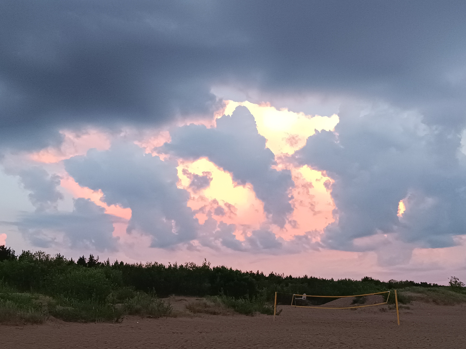 The image depicts a beach scene with a volleyball net set up on the sand in the foreground. In the middle ground, a dense line of trees forms the horizon. The sky above is filled with dramatic clouds, illuminated by the setting sun with hues of pink, yellow, and gray. The lighting suggests it is late evening. The scene is devoid of people. The specific location in the city of Riga, Latvia cannot be determined from the image.